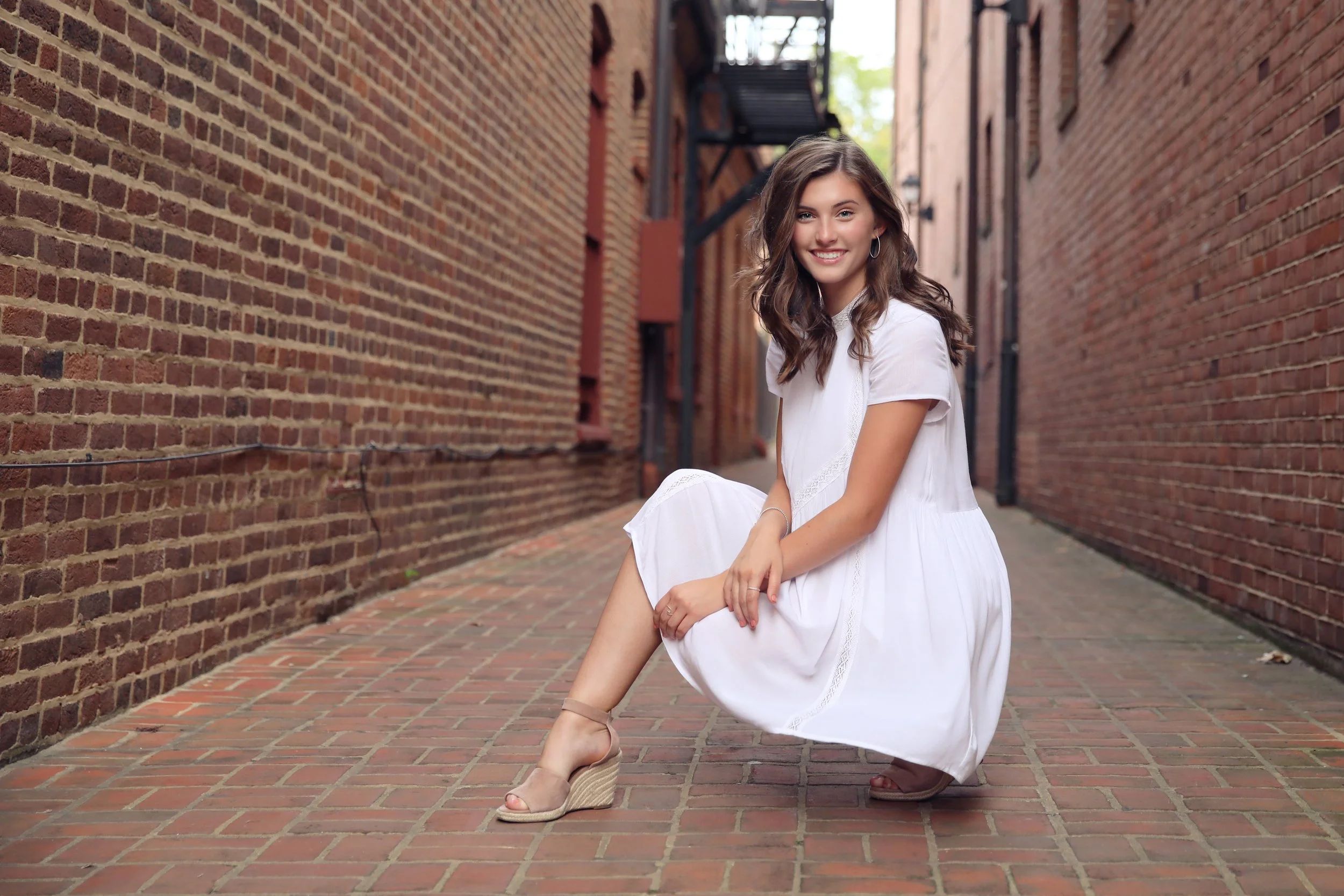 A young woman in a white dress and beige wedge sandals is squatting on a brick-paved alley between two brick buildings, smiling at the camera.