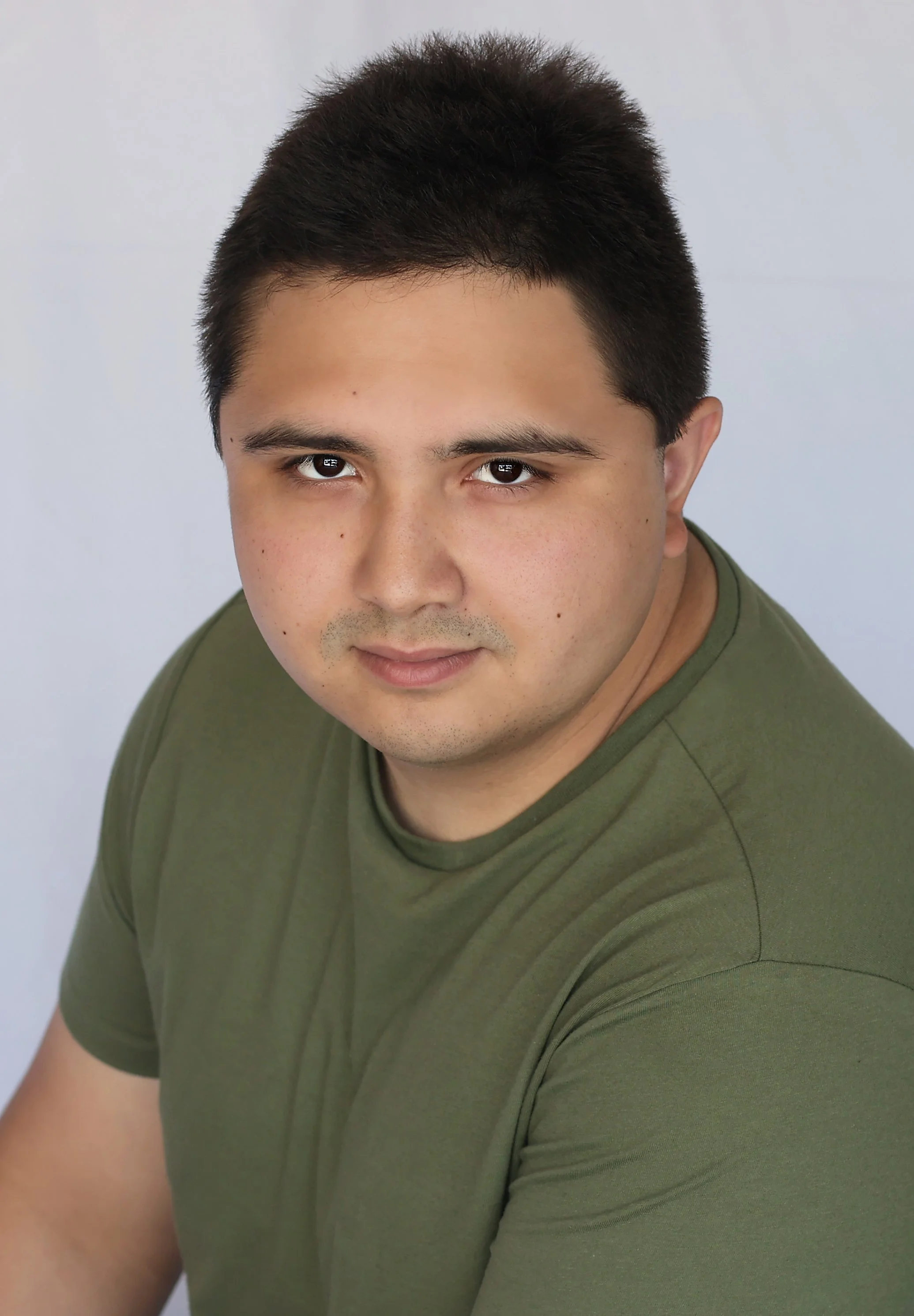 A young man with short dark hair, light skin, wearing an olive green t-shirt, looking directly at the camera against a neutral background.