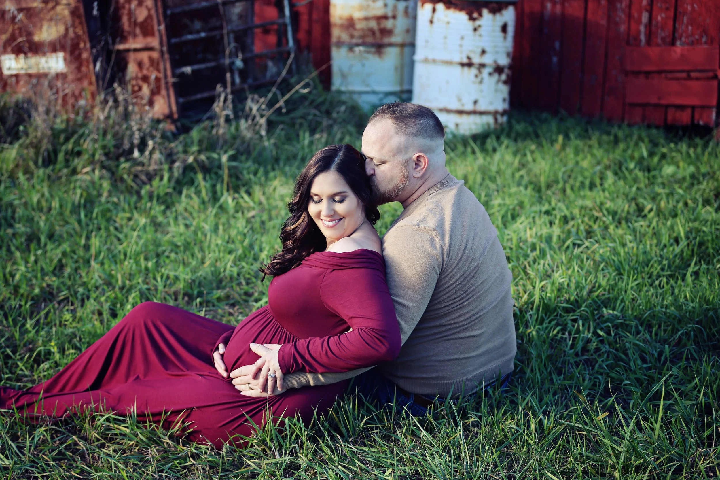 A pregnant woman in a long burgundy dress, sitting on the grass with a man, both smiling, with the man kissing her forehead. They are outdoors, with a red fence and some rustic objects in the background.