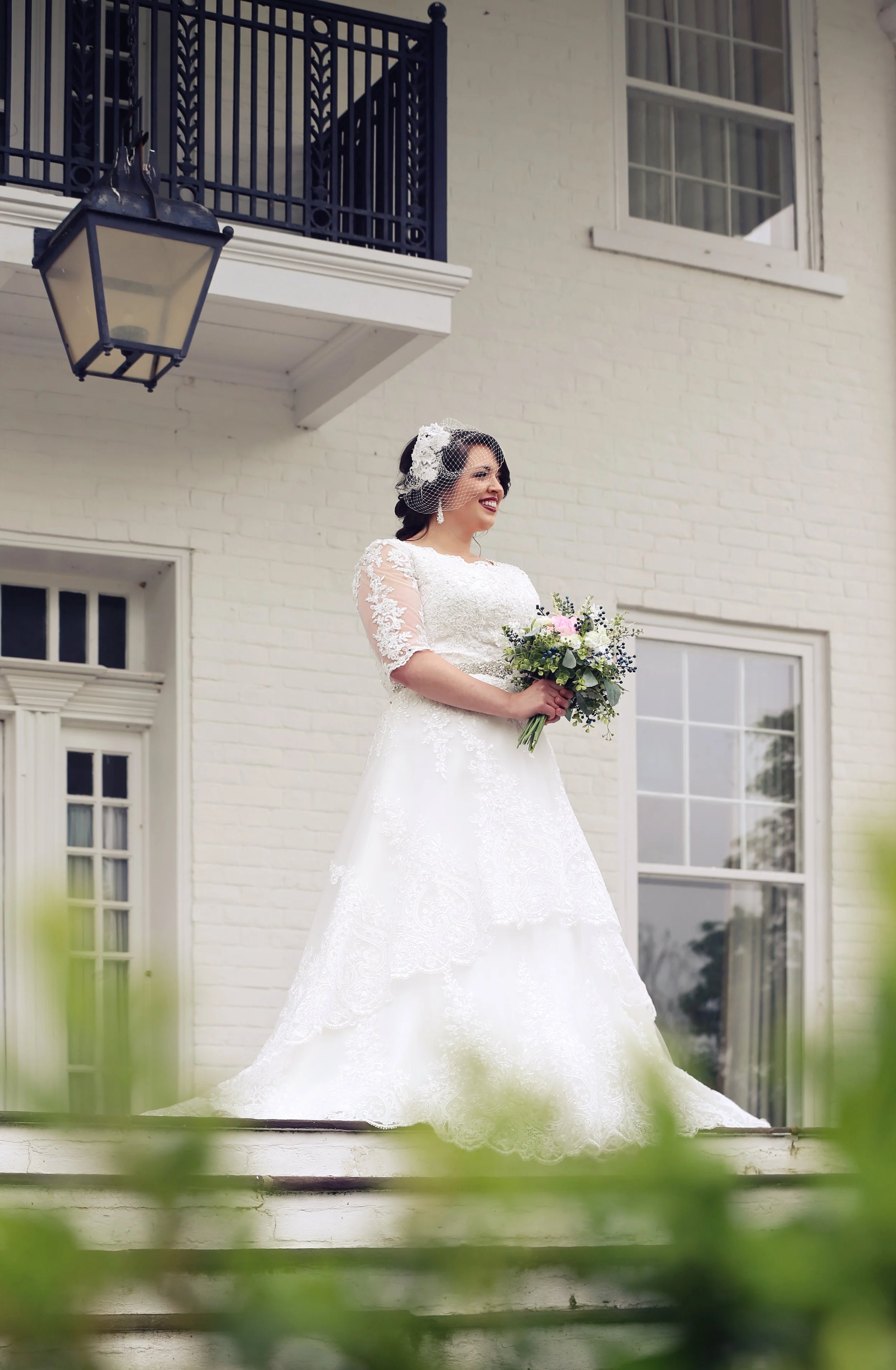A bride standing outdoors on a step in front of a white brick building, holding a bouquet, wearing a lace wedding dress with three-quarter sleeves, a birdcage veil, and smiling.