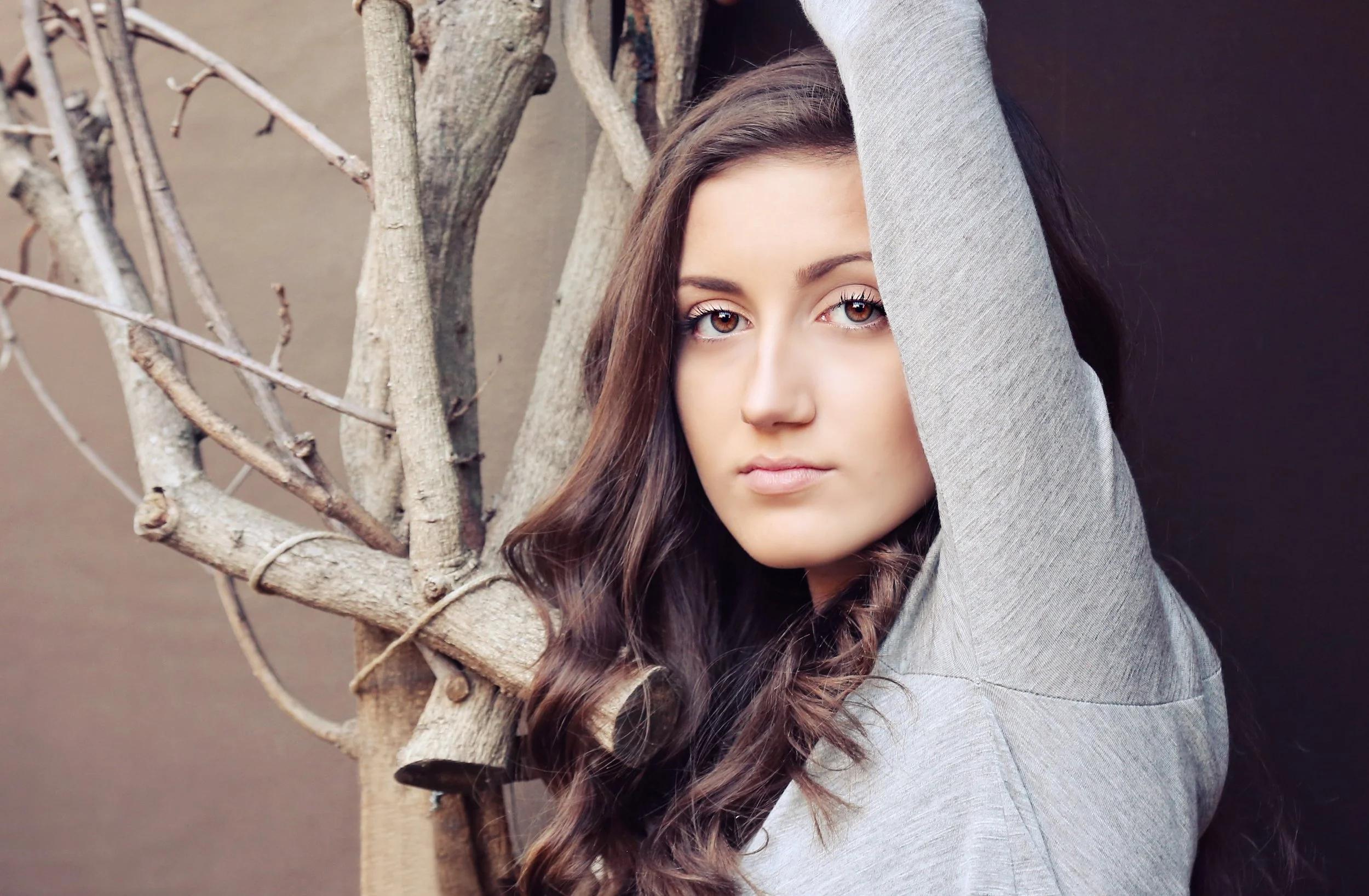 A young woman with brown hair and brown eyes leaning against a tree with bare branches, wearing a gray long-sleeve shirt.