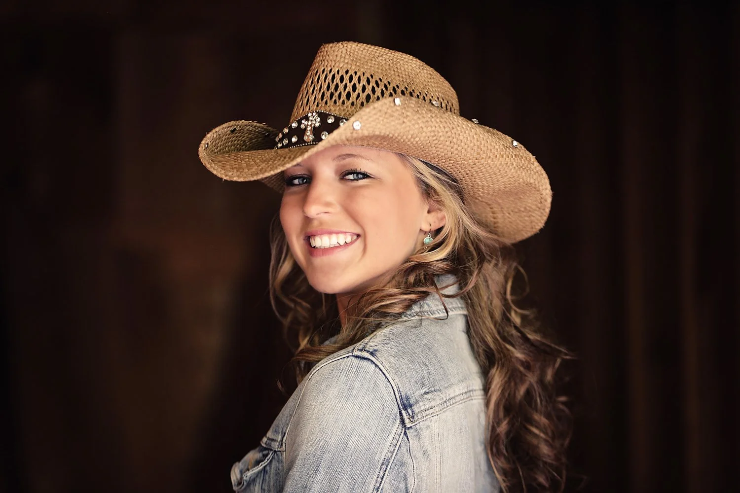 A young woman with curly hair wearing a straw cowboy hat with a black band and rhinestones, smiling and looking over her shoulder against a dark background.