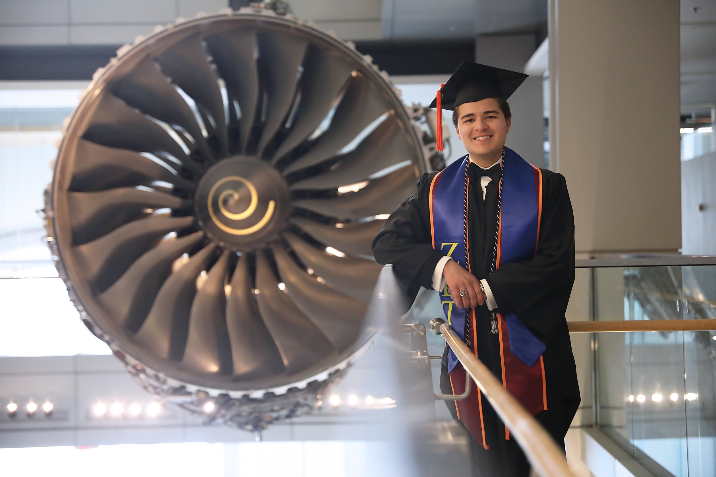 A young man in graduation attire, including a cap and gown, smiling and standing beside a large jet engine on display in a modern building.