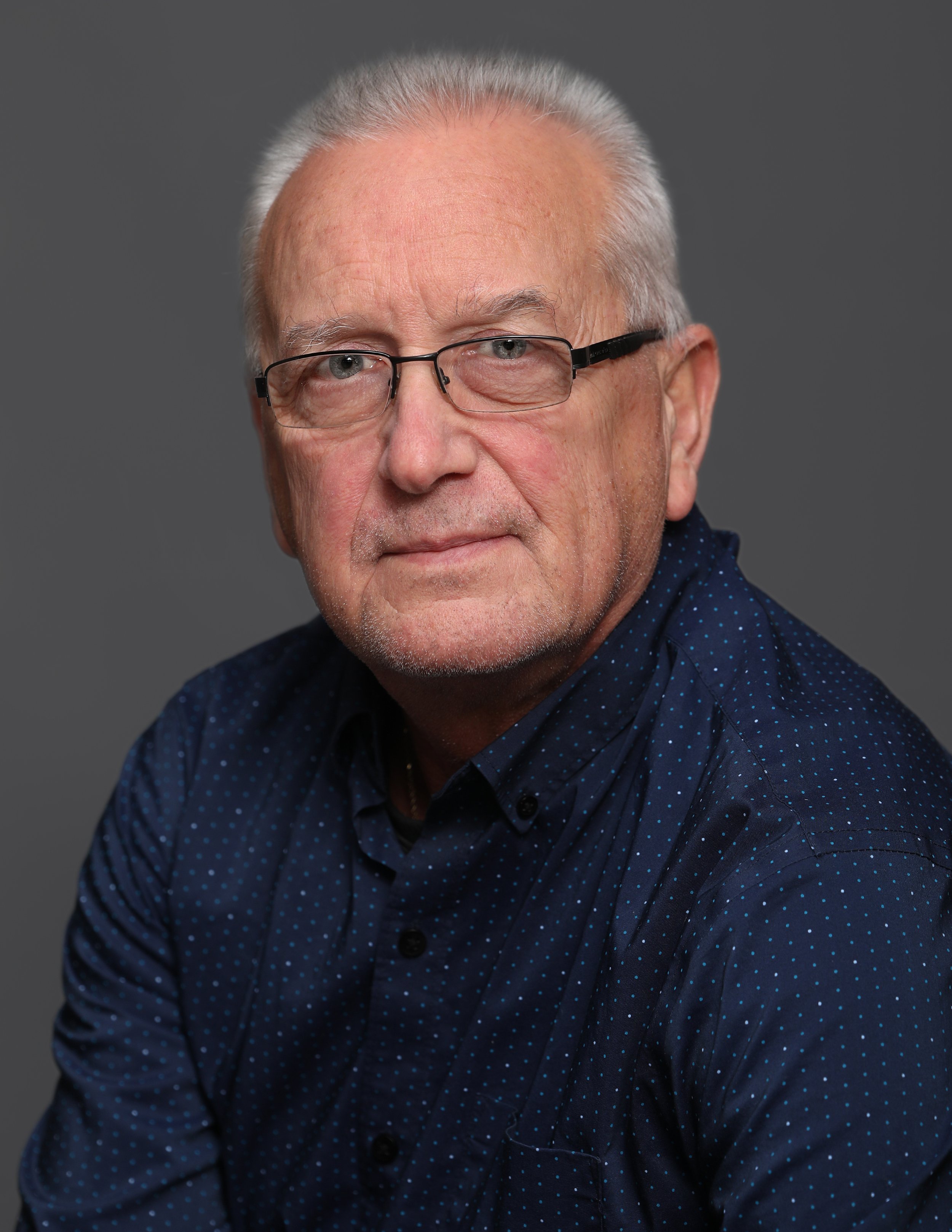 Portrait of an older man with gray hair, glasses, and a navy blue dotted shirt against a gray background.