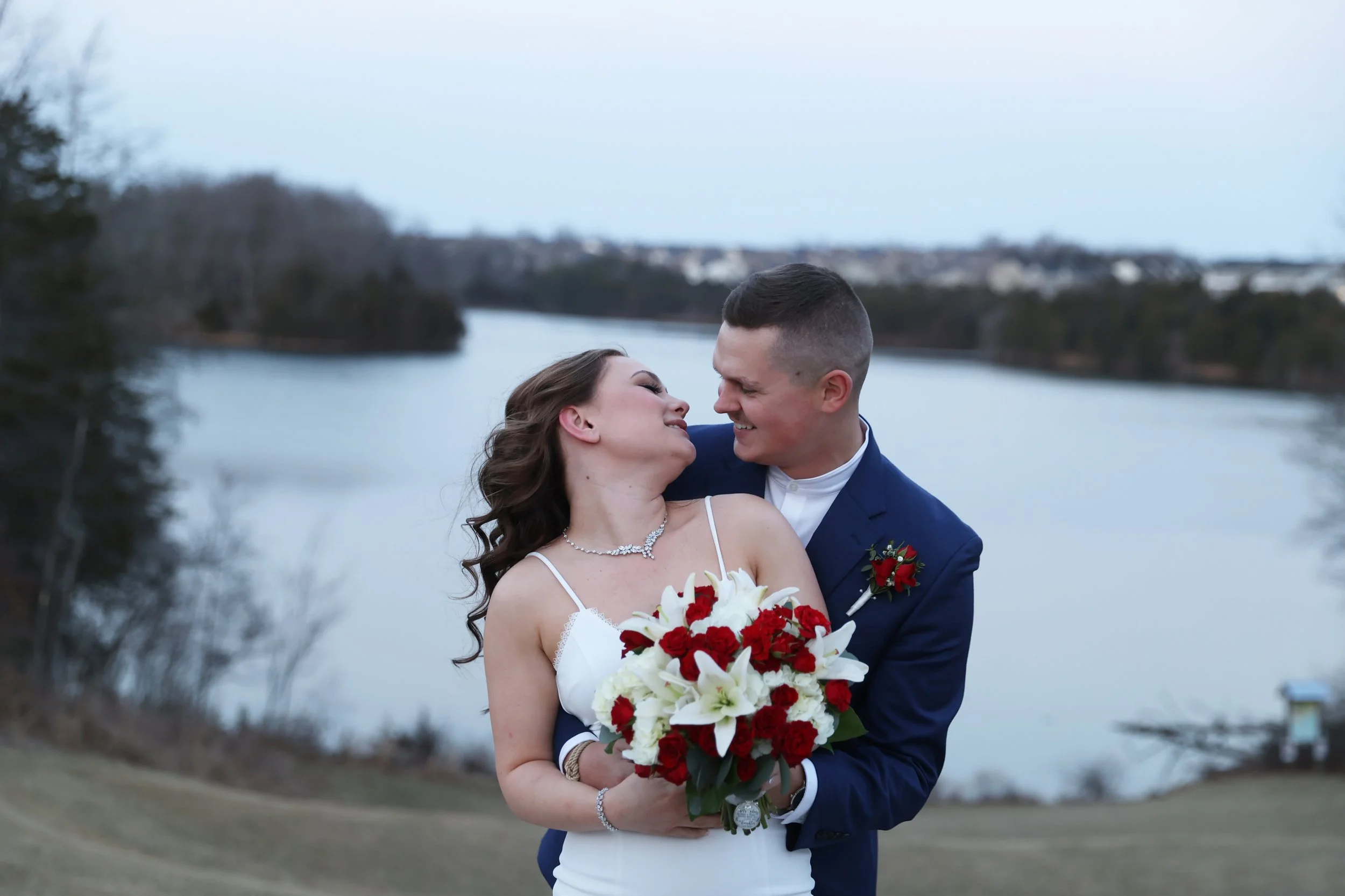 A newlywed couple embracing outdoors near a river, smiling at each other. The bride is holding a bouquet of white lilies and red roses, wearing a white wedding dress, and the groom is in a navy suit with a red boutonniere. The background features a river and overcast sky.