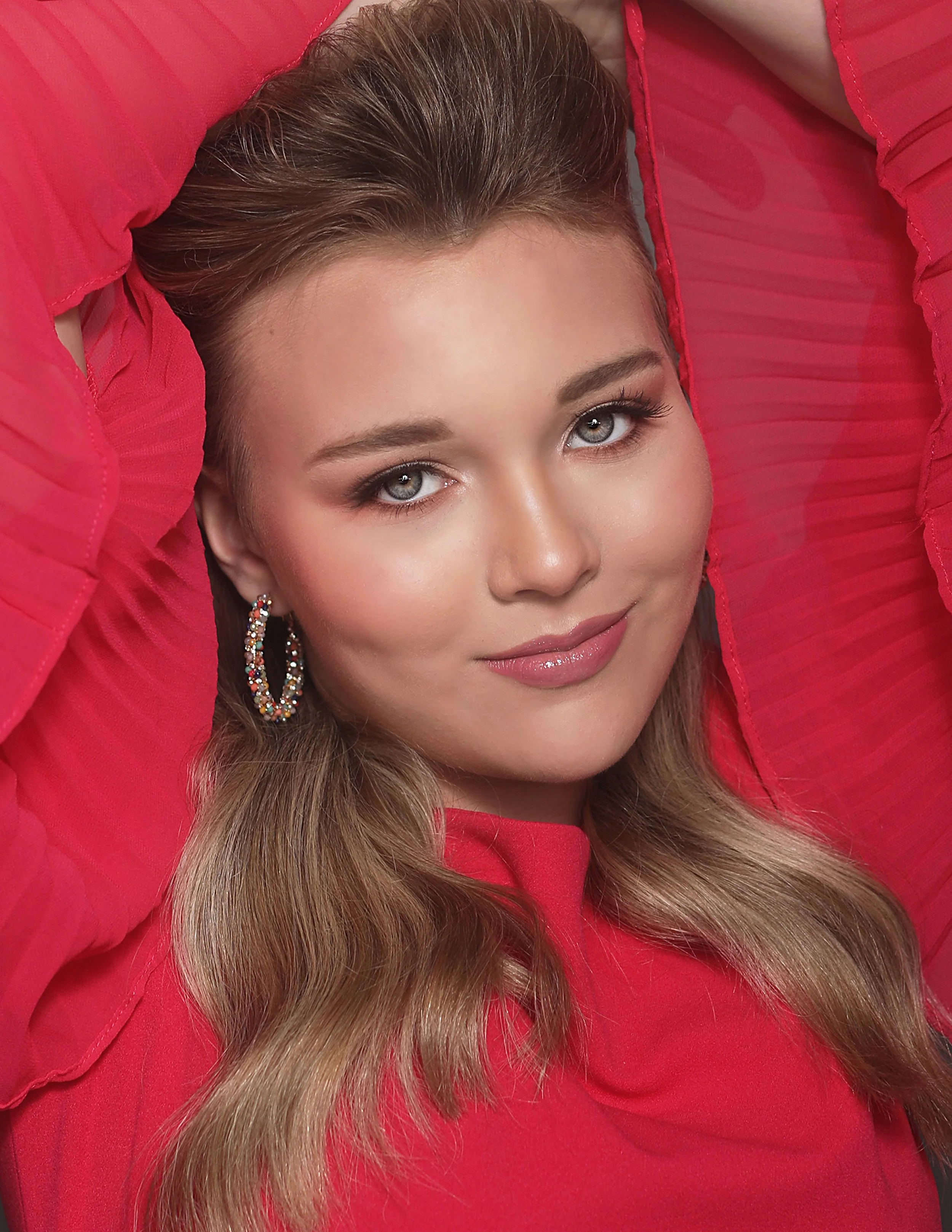 Close-up portrait of a woman with long wavy blonde hair, wearing hoop earrings and a red top, lying against a red background, with her arms positioned around her head.