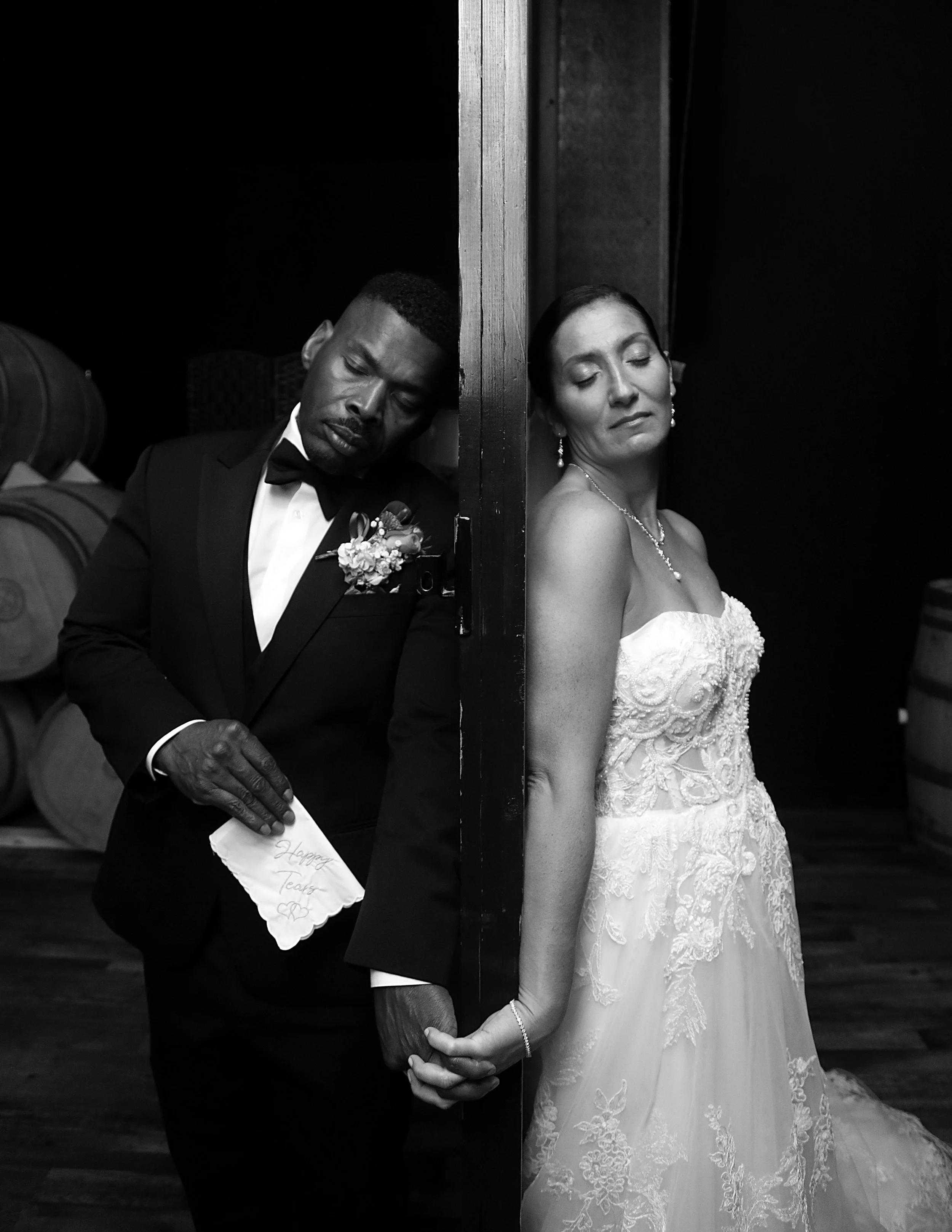 A black and white photo of a bride and groom holding hands, separated by a vertical wooden post. The groom is in a tuxedo with a boutonniere, and the bride is in a lace wedding gown with jewelry. They appear to be standing in a winery or cellar with wine barrels in the background.