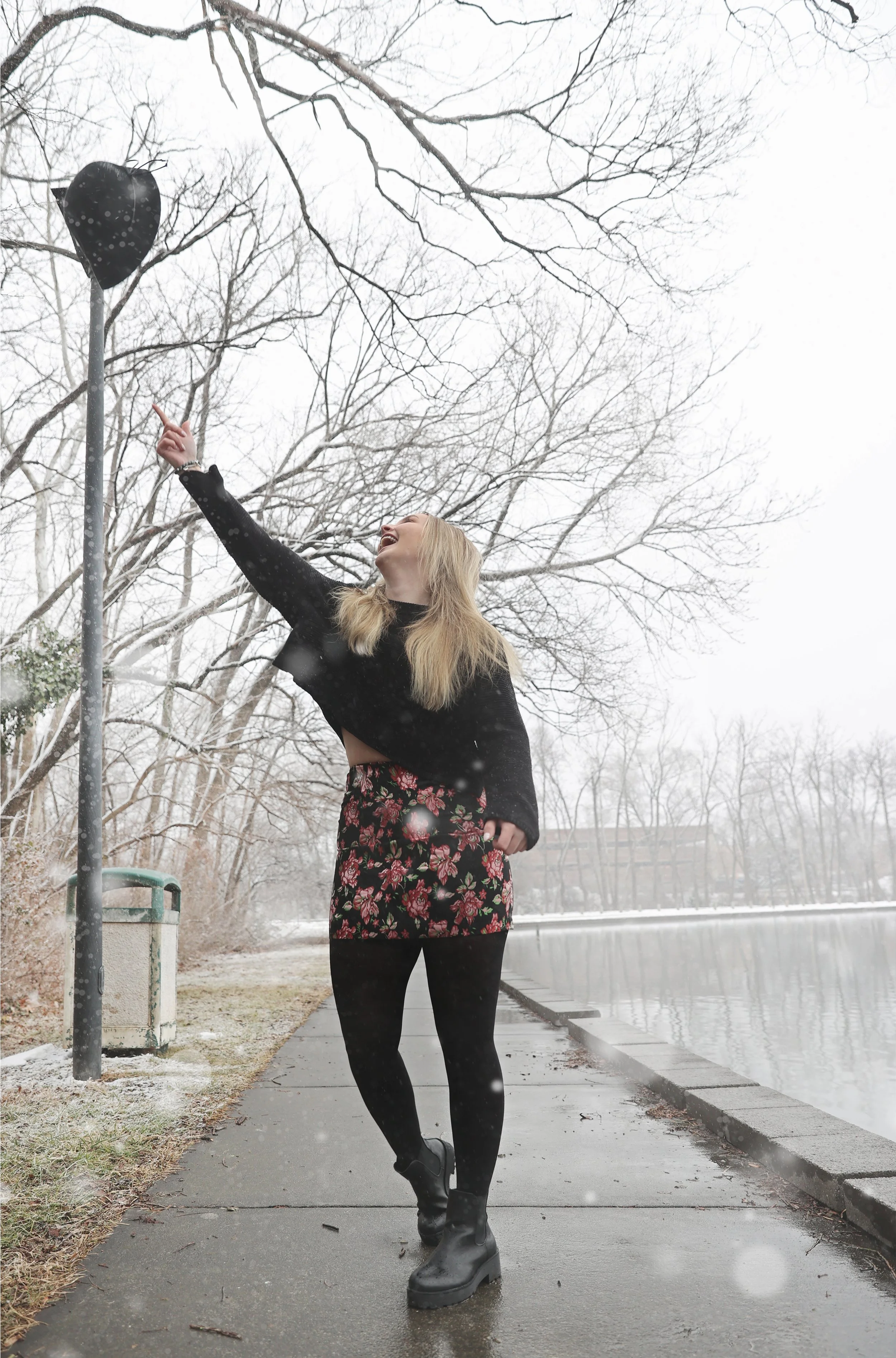 A woman in a black sweater, floral skirt, and black tights standing on a sidewalk near a body of water on a winter day, pointing and smiling at a heart-shaped balloon attached to a pole.