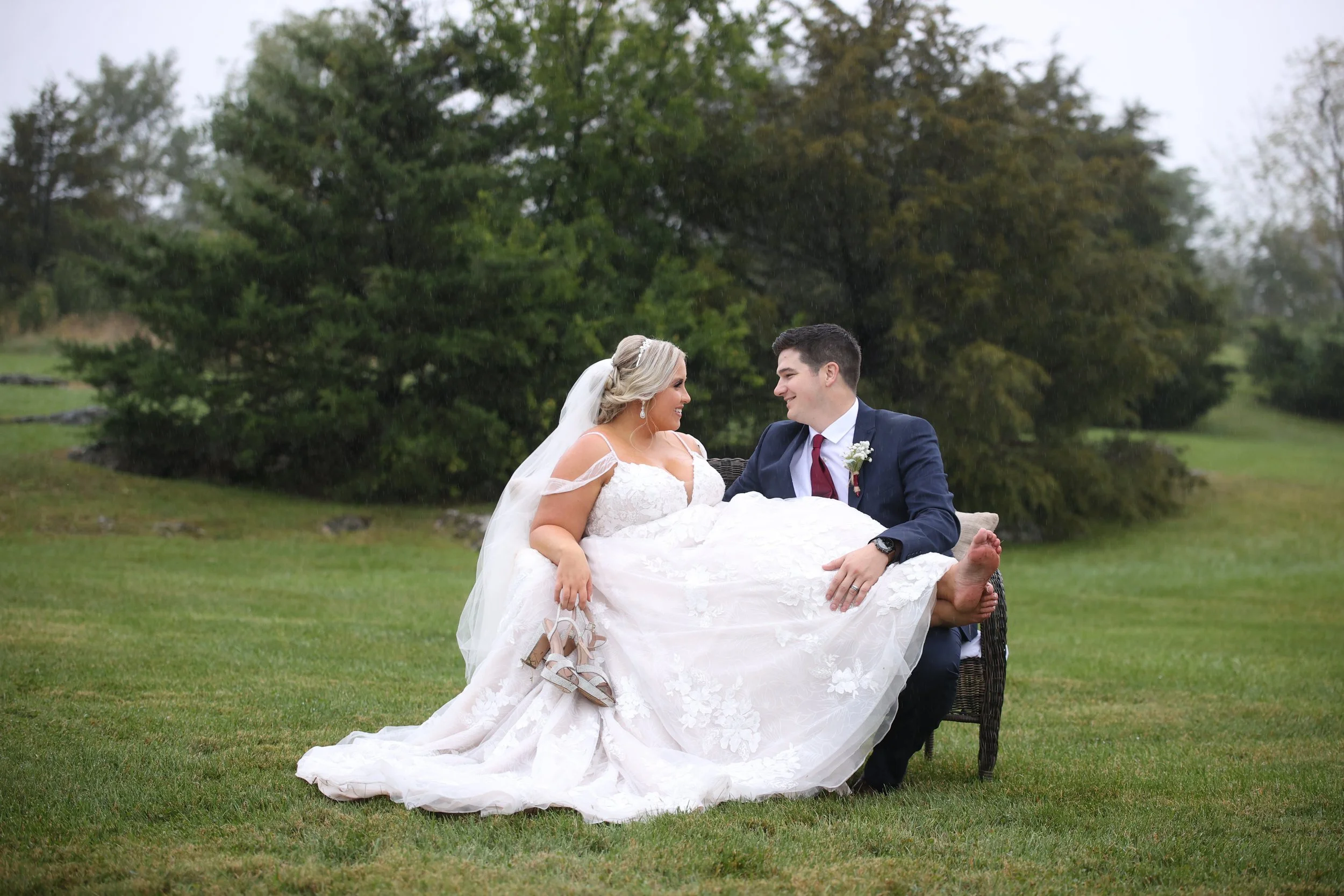 A bride and groom sitting outdoors on a bench, smiling at each other. The bride is holding her shoes in her hand, and the groom is wearing a dark suit with a red tie. The scene is set in a grassy area with trees in the background.