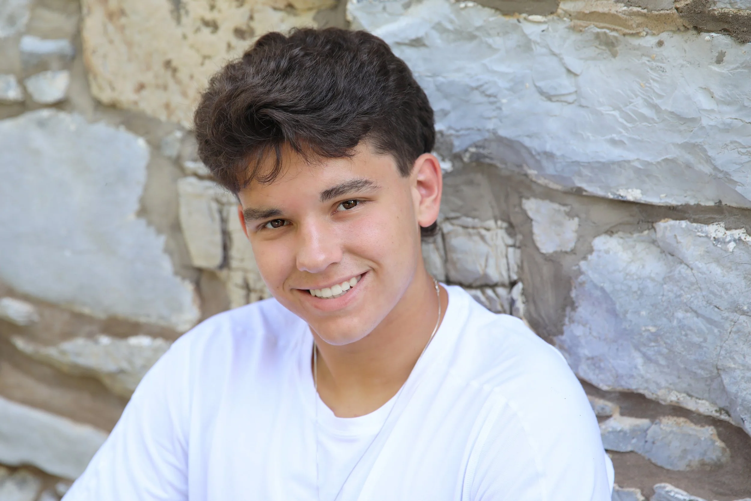 Young man with dark hair and a white shirt smiling against a stone wall background.