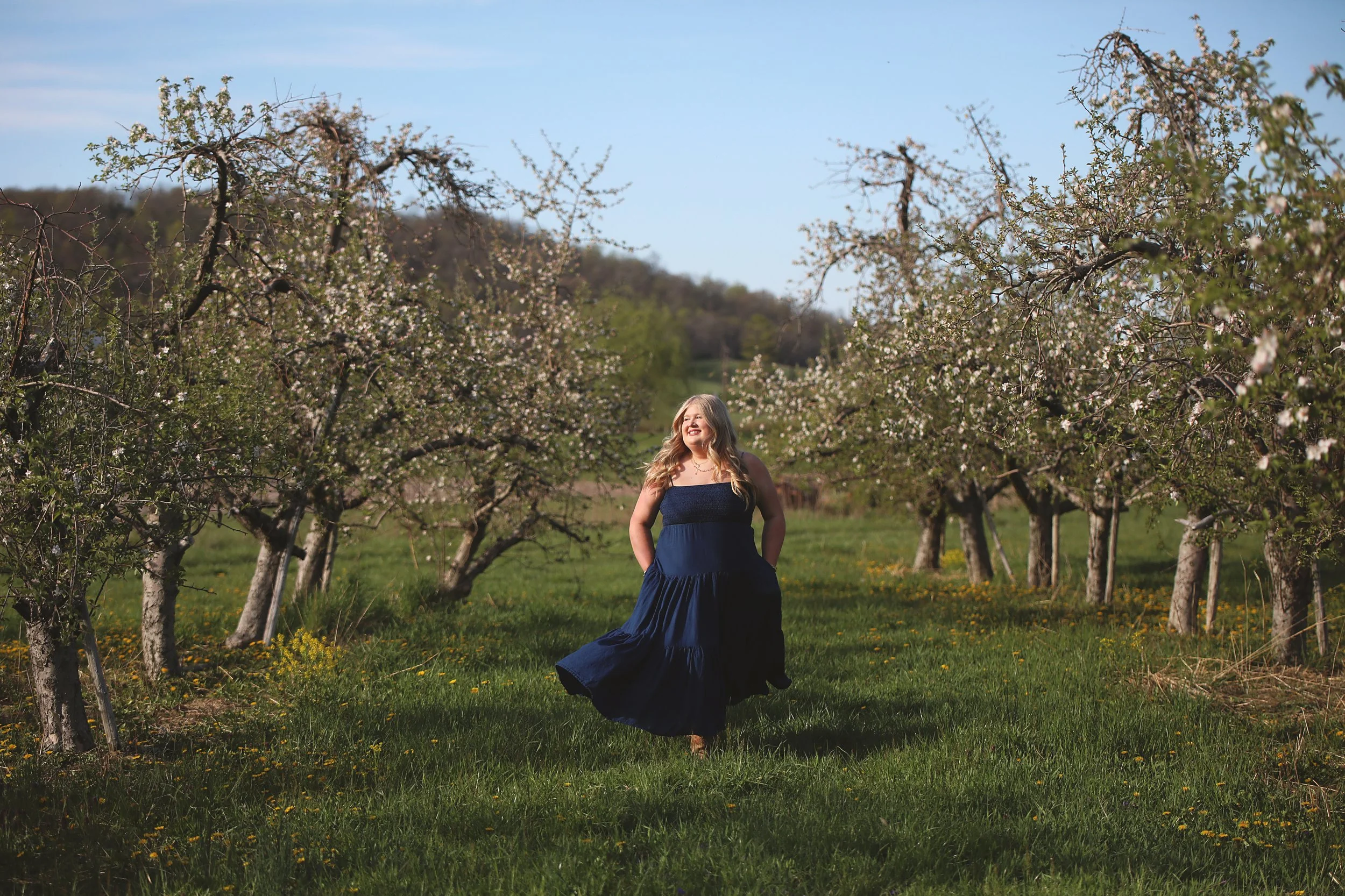 A woman in a navy blue dress walking through an orchard of blossoming fruit trees, smiling and enjoying the sunny day.