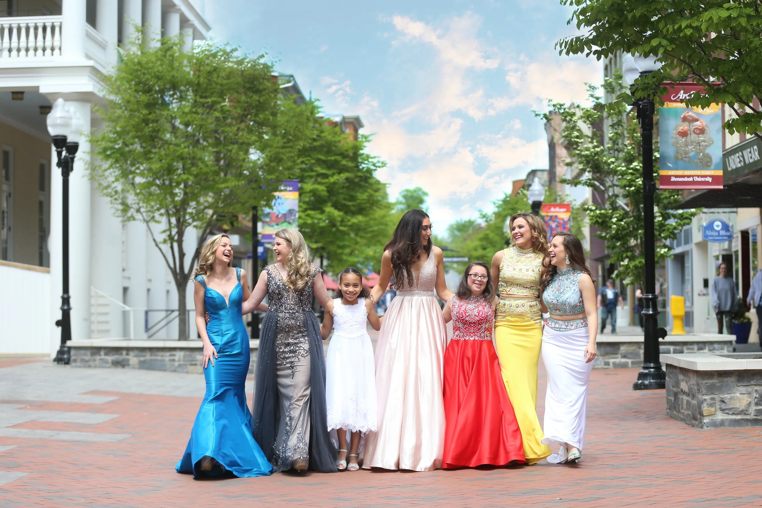 Group of seven young women smiling and walking together on a brick sidewalk in a city street, dressed in formal evening gowns of various colors, with trees and shops lining the street.