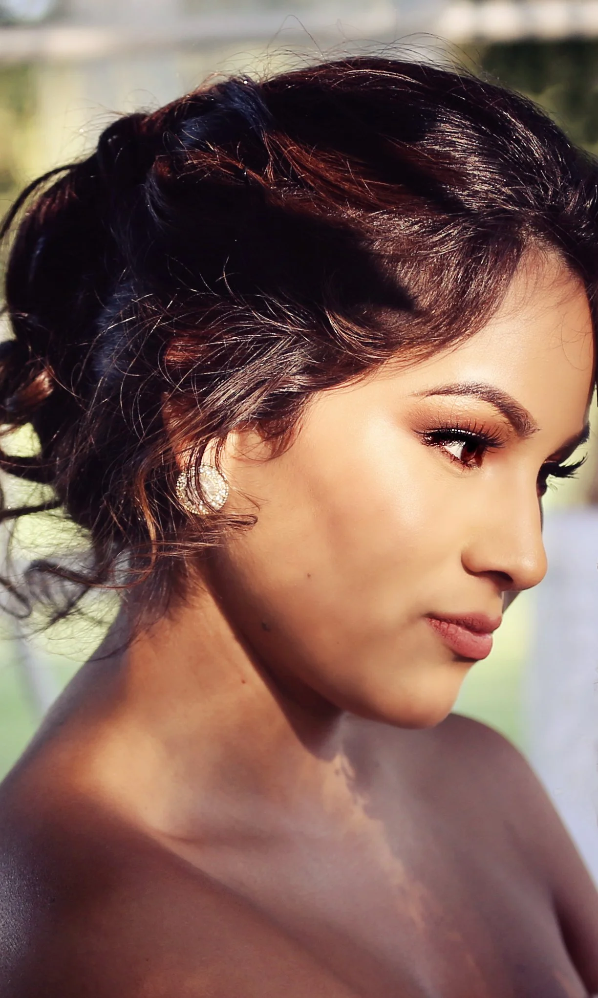 Close-up of a woman with styled dark hair, wearing sparkly earrings, and detailed makeup, outdoors with natural light.