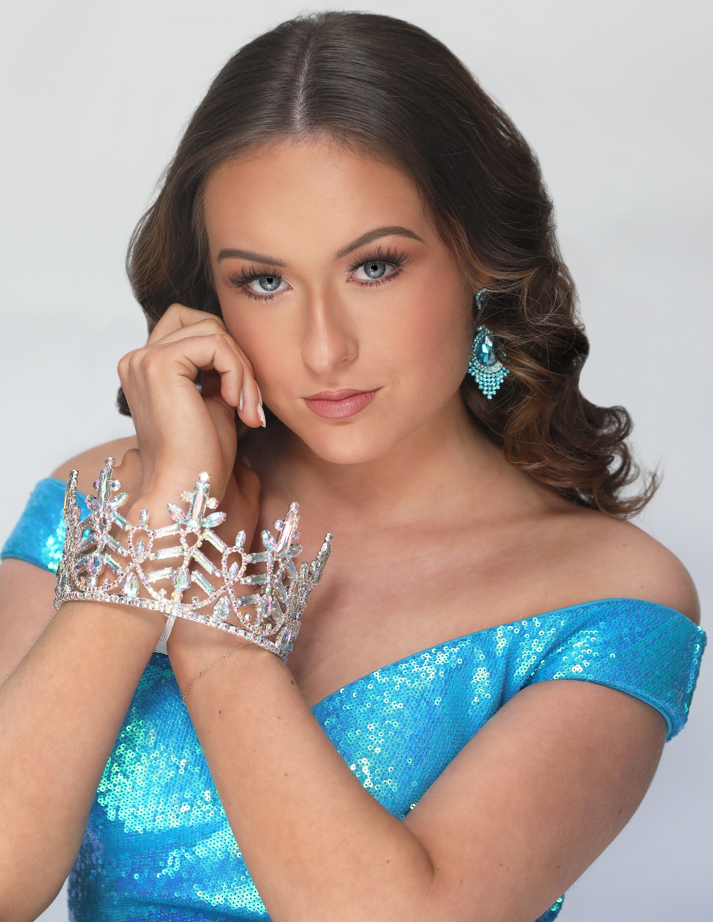 A woman with brown, wavy hair and blue eyes wearing a sparkly blue dress, large blue earrings, a crown-shaped bracelet, and a delicate necklace, looking at the camera with her hand resting on her face against a plain white background.