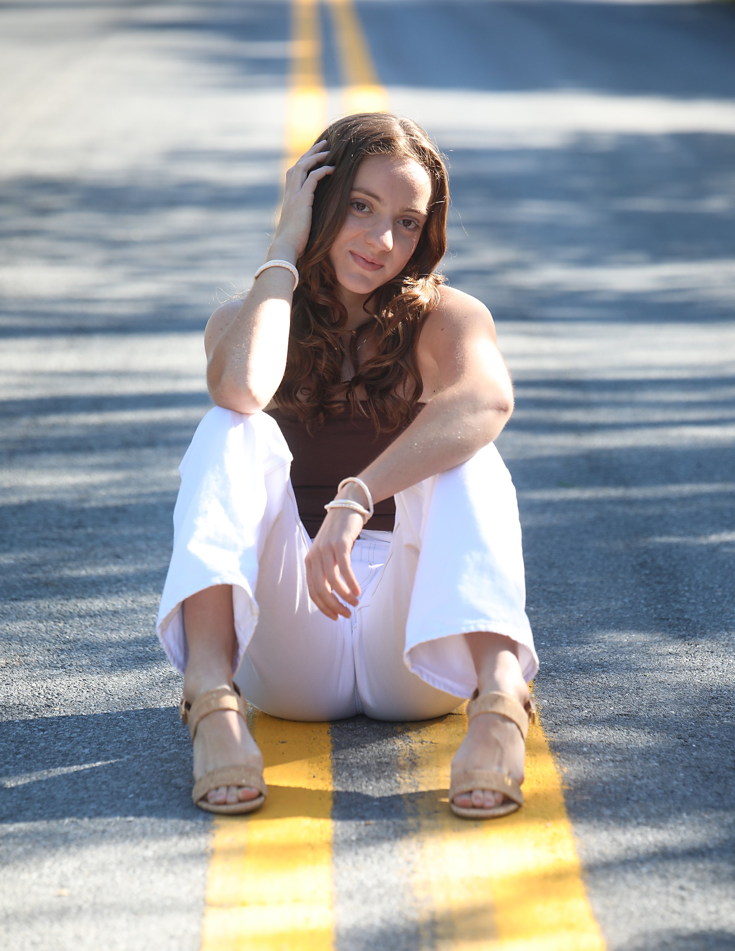 A young woman with long, curly brown hair sitting on an empty road with yellow lines, wearing a brown tank top, white pants, and beige heels, looking at the camera with a gentle smile.