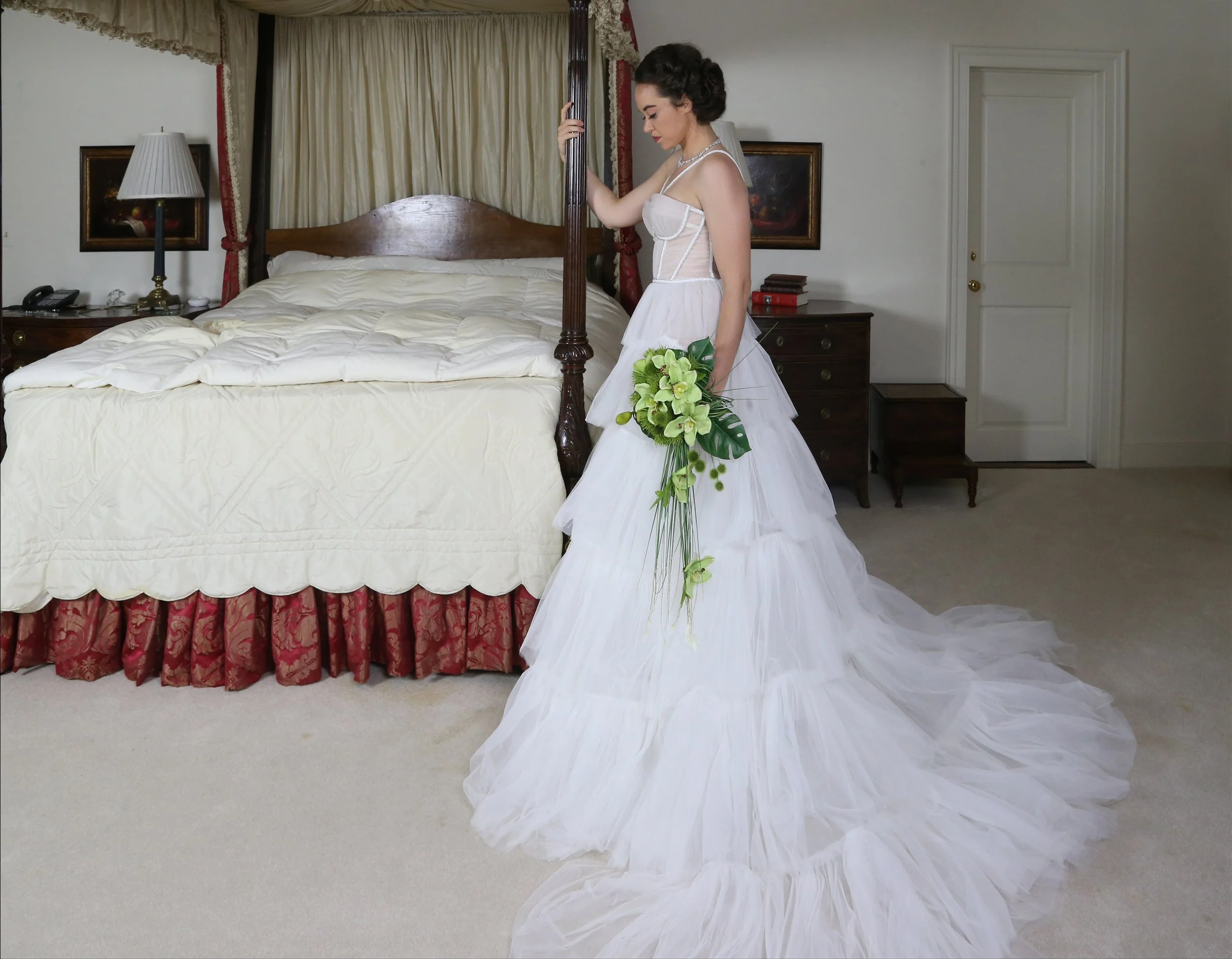 A bride in a white wedding gown holding a bouquet of green flowers and leaves, standing next to a large bed with a cream-colored bedspread and red and gold curtains in a hotel room.