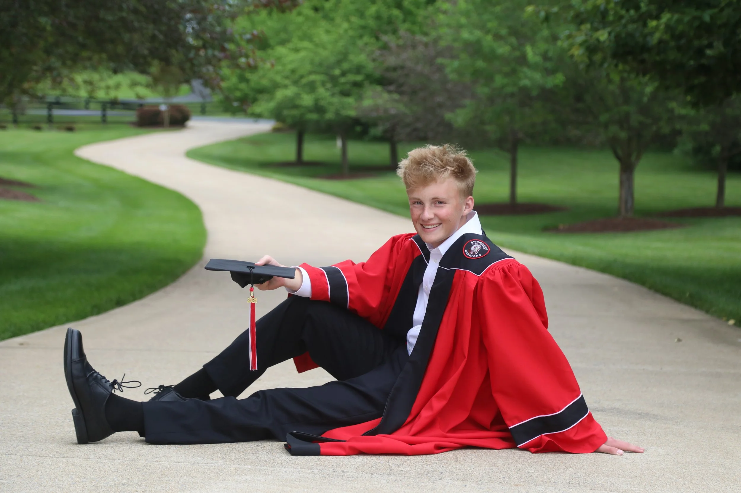 A young man in a graduation cap and gown sitting on a sidewalk, smiling at the camera, with a lush green park in the background.