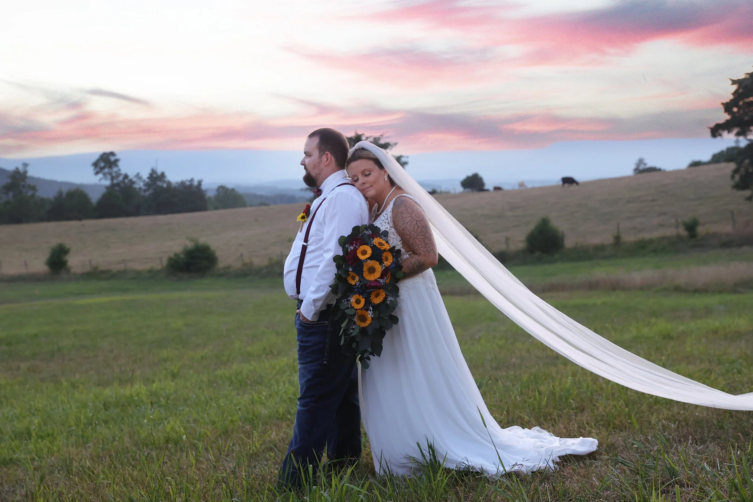 Bride and groom standing in a grassy field during sunset, with the bride holding a sunflower bouquet, the bride leaning on the groom's shoulder, and a flowing veil.