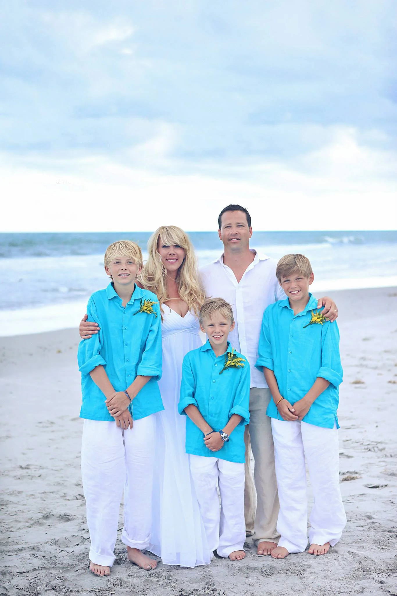 Family of five standing on the beach with the ocean in the background, dressed in white and blue, smiling at the camera.