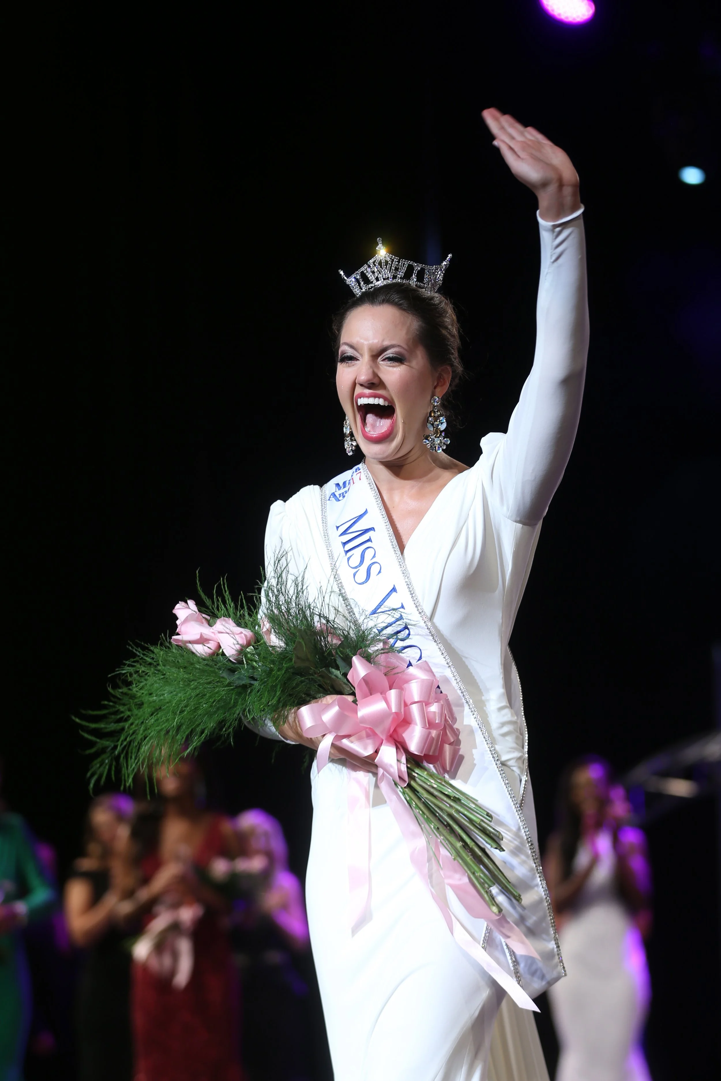 A woman wearing a white dress, a crown, and a sash that says "Miss Venezuela" is holding a bouquet of pink roses and celebrating on stage.