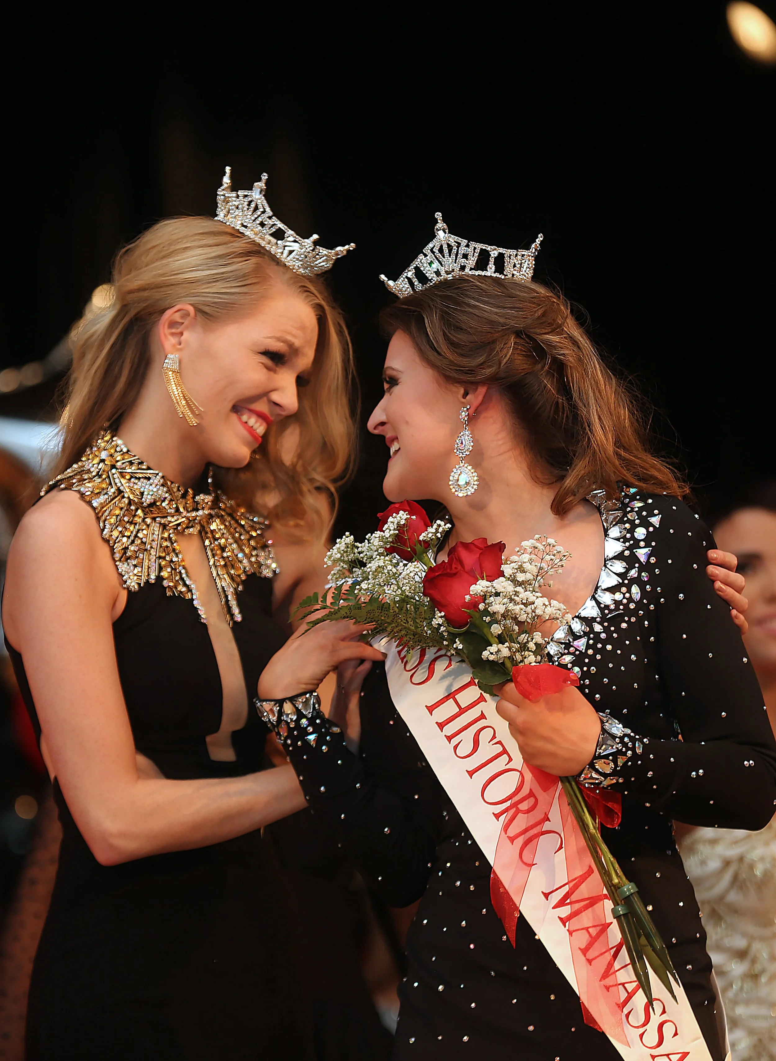 Two women wearing crowns and tiaras smiling at each other. One woman is holding a bouquet of red roses and wearing a sash that reads 'Miss Horticultural.' They are dressed in glamorous outfits with jewelry and are at a celebratory event.