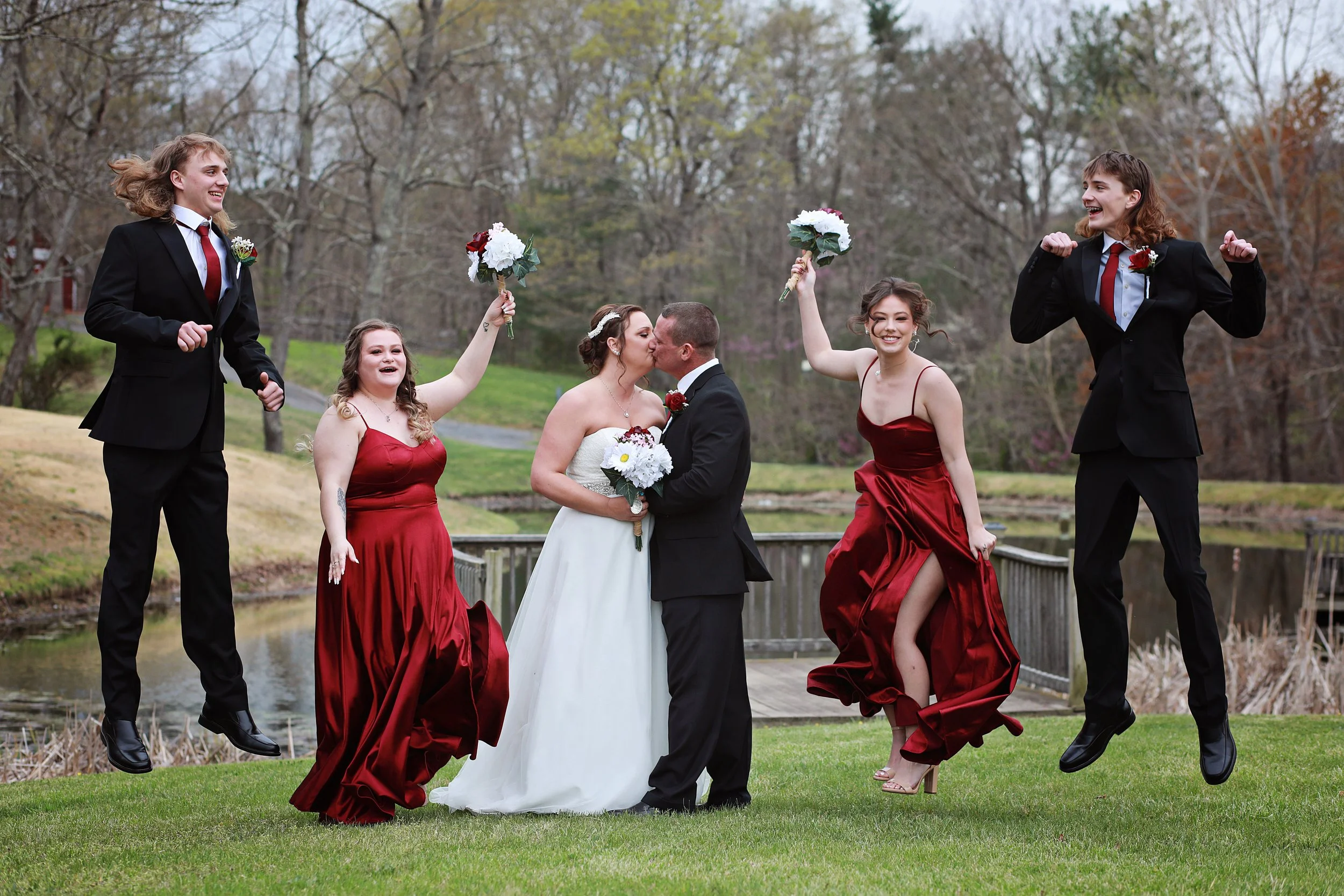 A wedding party celebrating outdoors with the bride and groom kissing in the center, surrounded by bridesmaids and groomsmen, some of whom are jumping and holding bouquets, with a pond and trees in the background.