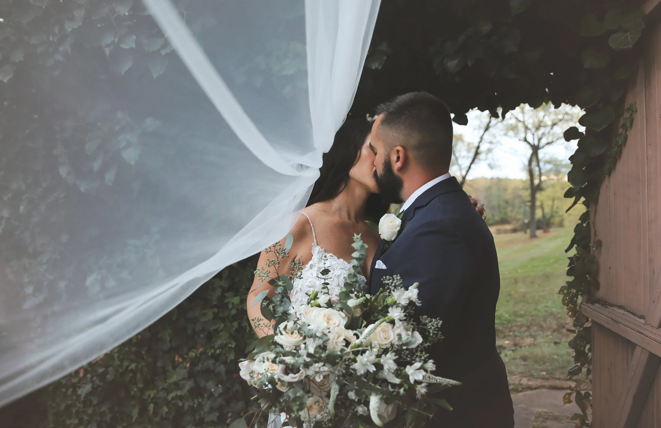 A bride and groom kissing outdoors under a sheer curtain, with greenery and trees in the background. The bride is holding a large floral bouquet, and she is wearing a white lace wedding dress. The groom is in a navy suit with a white shirt and boutonniere.
