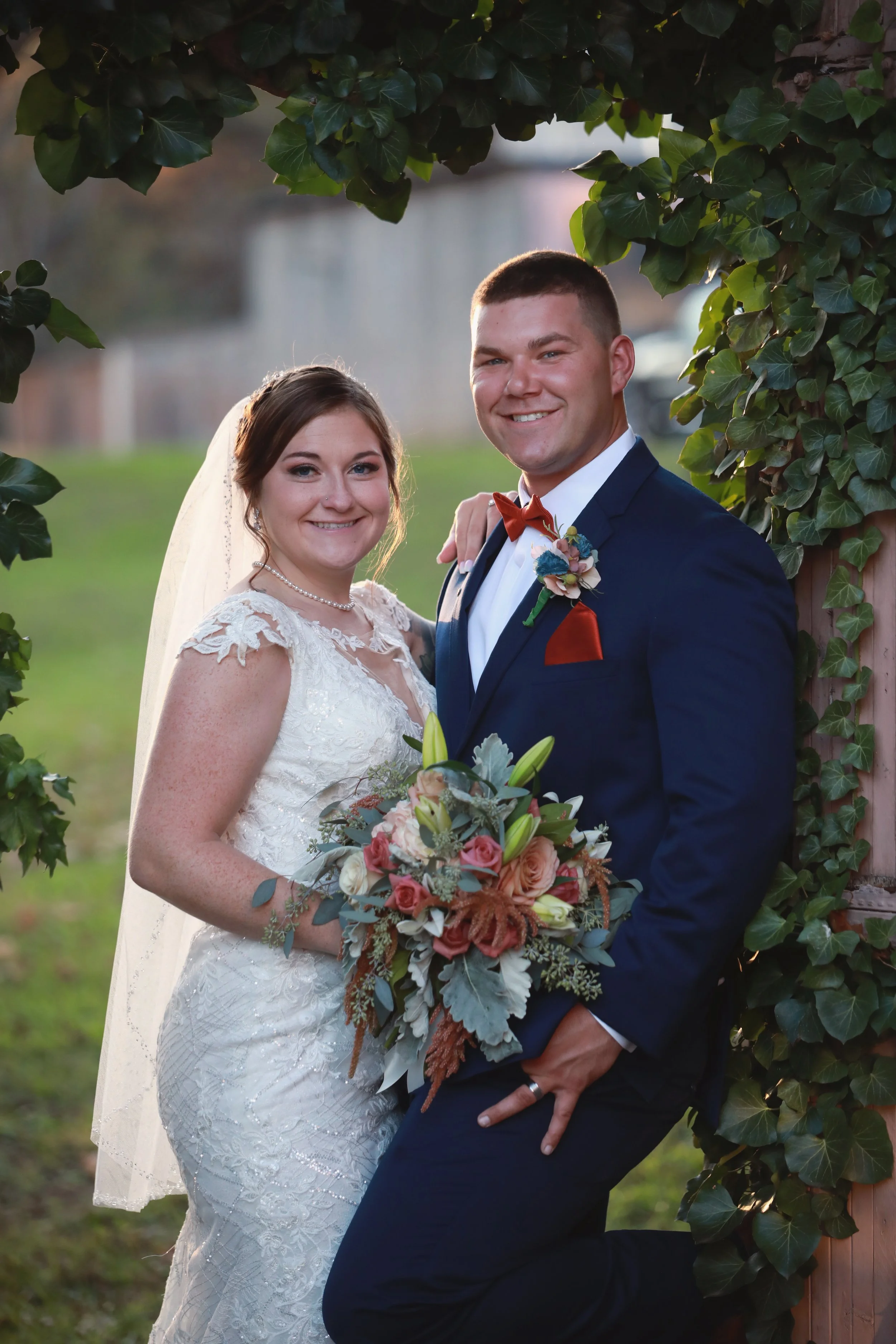A bride and groom sharing a pose outdoors, with greenery and ivy framing them, during a wedding celebration.