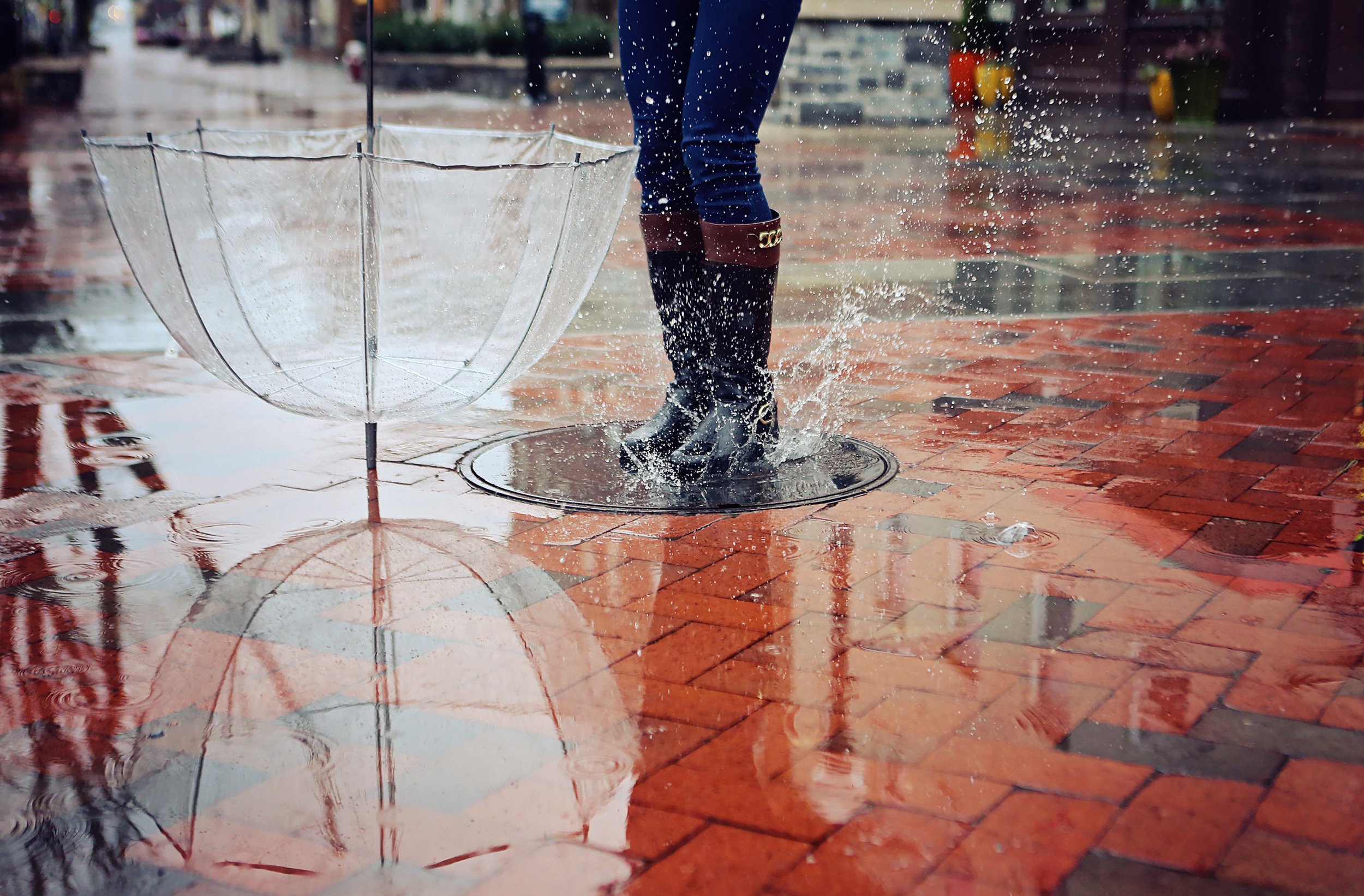 Person standing in a rain puddle with a clear umbrella, wearing rain boots and jeans, on a wet brick sidewalk.