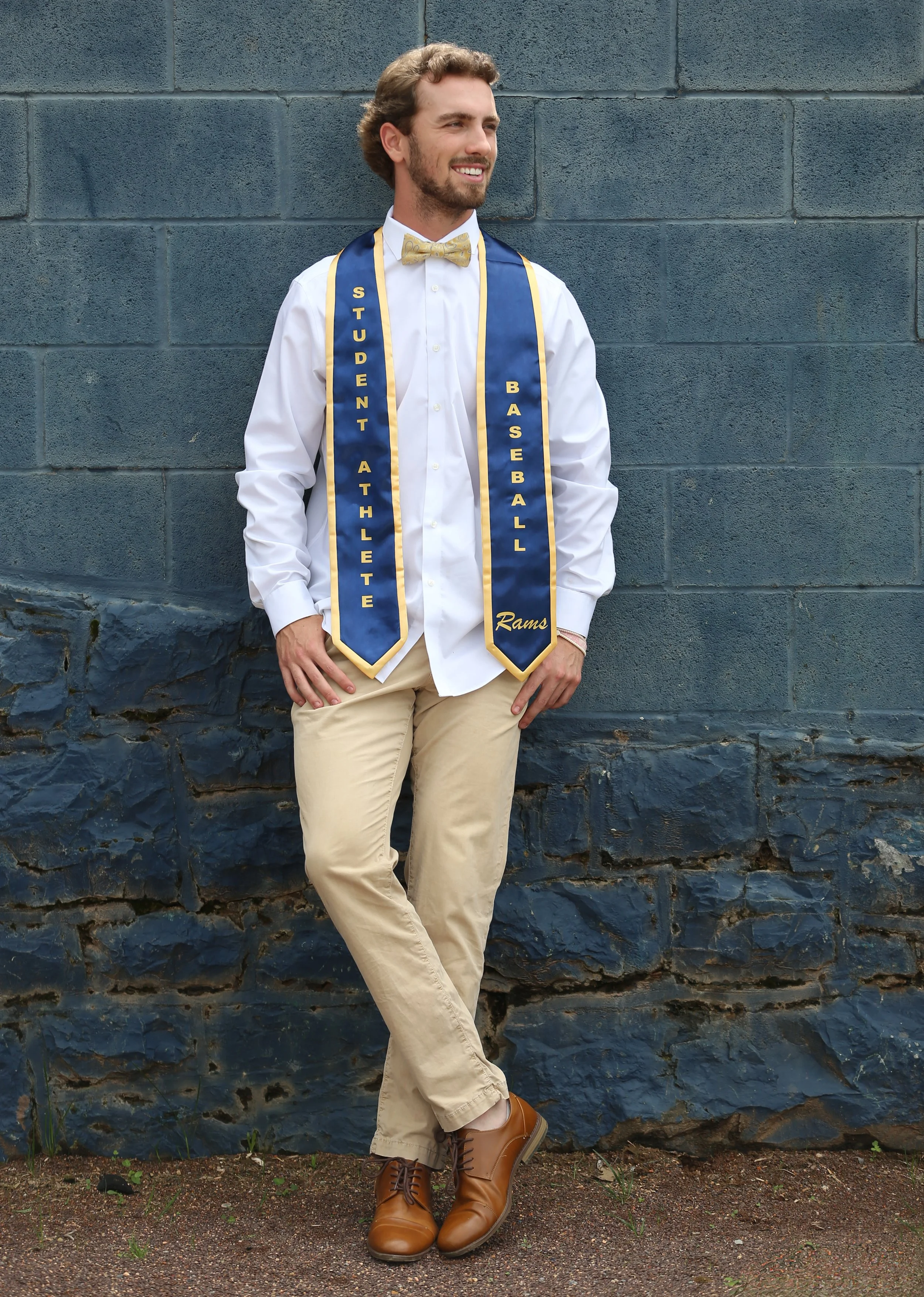 A man dressed in formal attire including a white shirt with a gold bow tie, tan pants, and brown shoes, standing against a gray brick wall. He is wearing a sash that reads 'STUDENT ATHLETE BASEBALL Rams'.