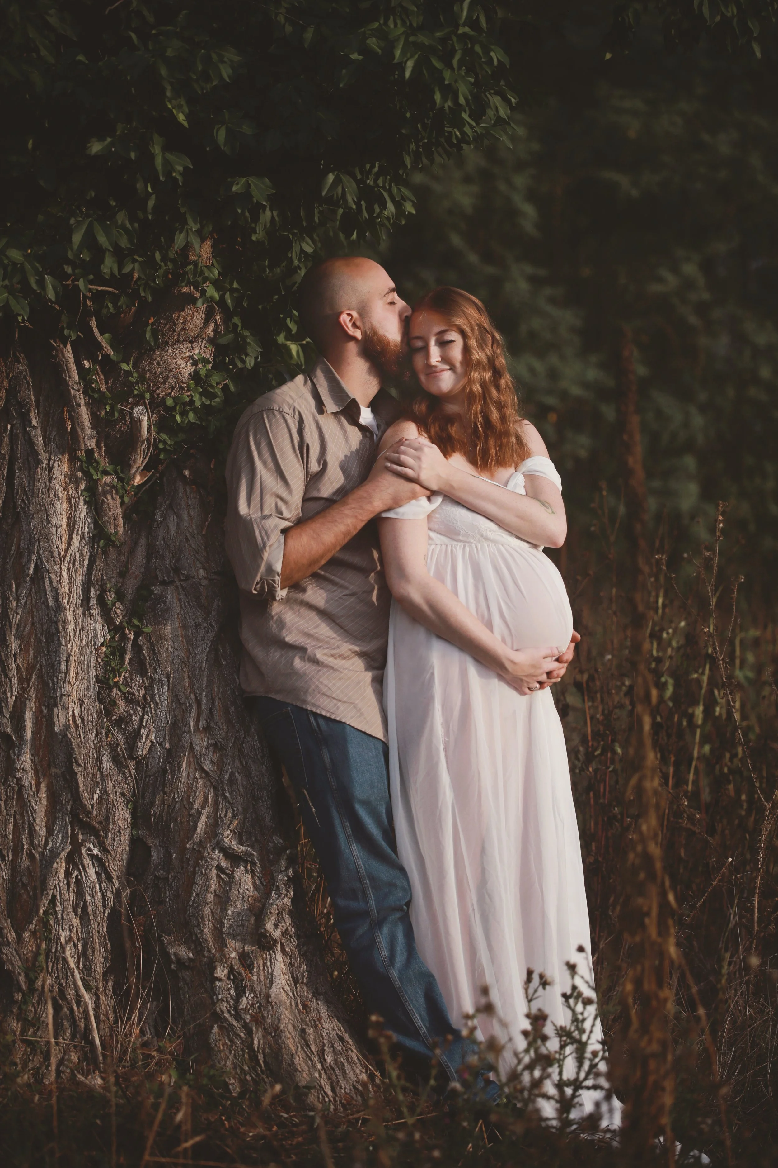 A pregnant woman with red hair in a white dress standing outdoors next to a man with a beard and bald head wearing a beige shirt and jeans, showing affection and embracing her while she gently holds her belly, with trees and foliage in the background.