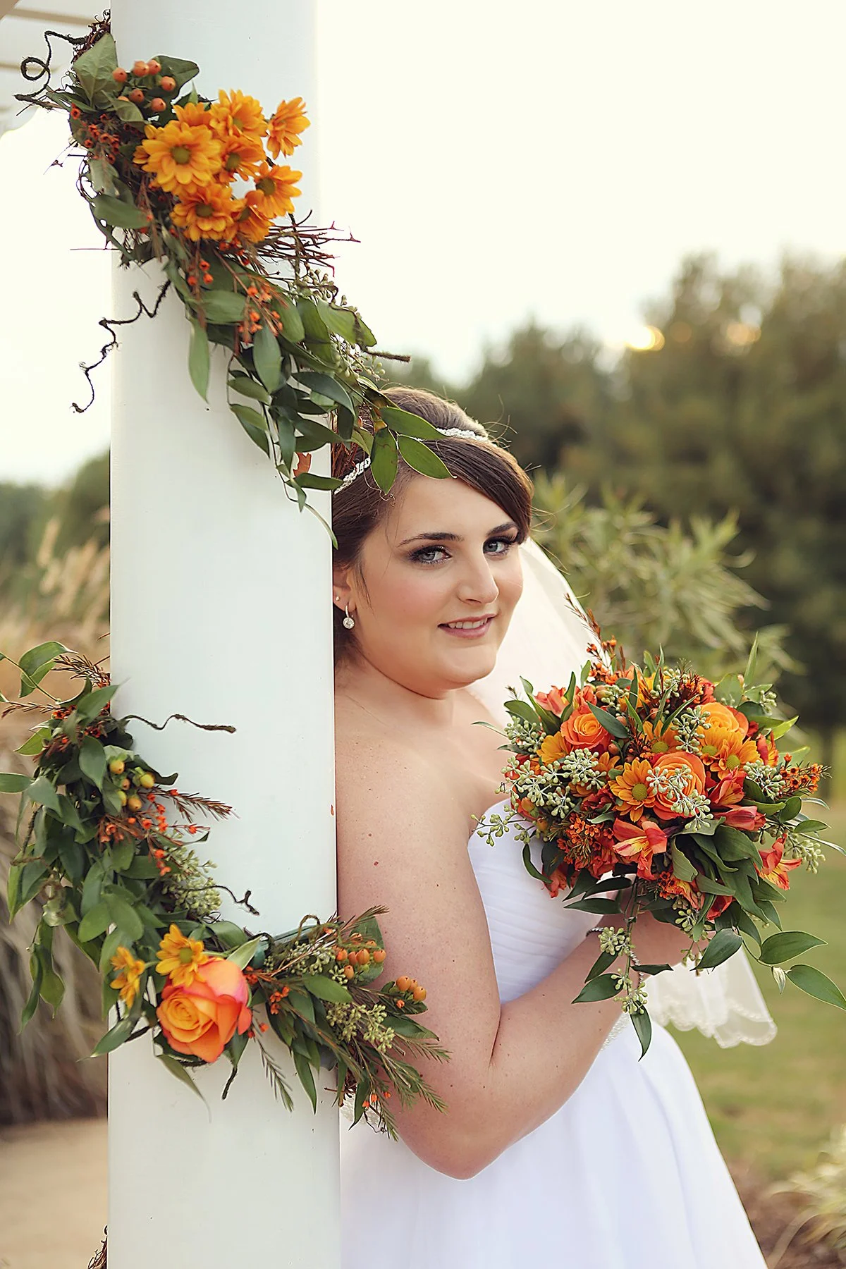 A bride peeks out from behind a white post decorated with orange and yellow flowers and green leaves, holding a matching bouquet outdoors during sunset.