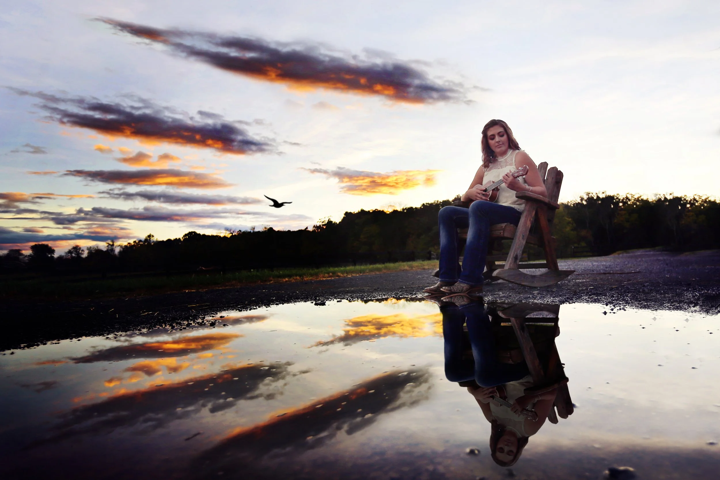 A girl sitting on a wooden rocking chair outdoors at sunset, playing a guitar, with a large puddle reflecting the sky and her figure, and a bird flying in the distance.