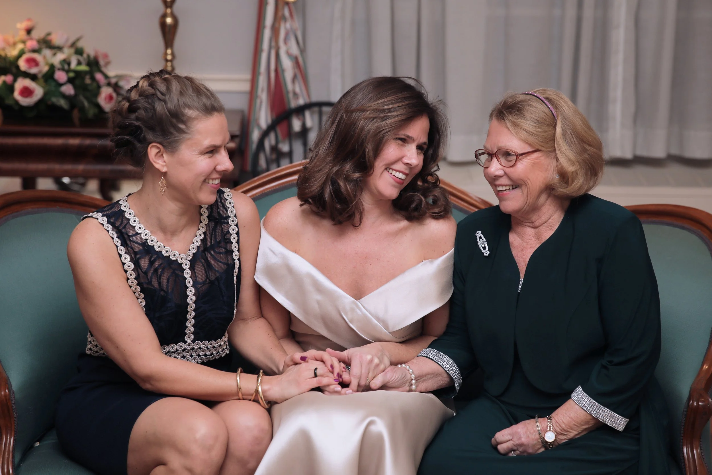 Three women sitting on a green couch, laughing and holding hands, in a decorated room, possibly at a wedding or celebration.