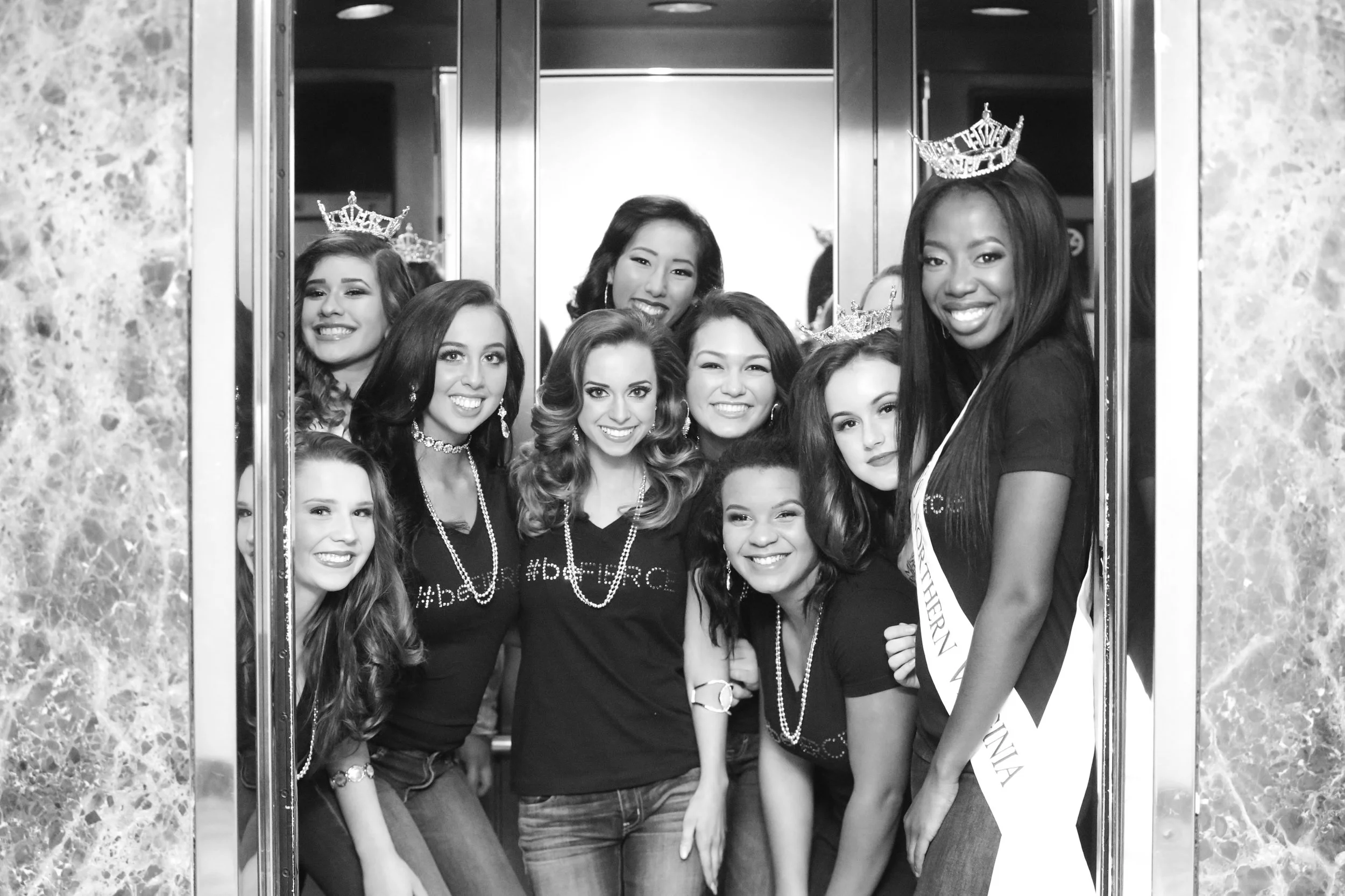 A group of young women wearing crowns and sashes, posing inside an elevator. They are smiling and posing for the photo, celebrating a pageant or similar event.