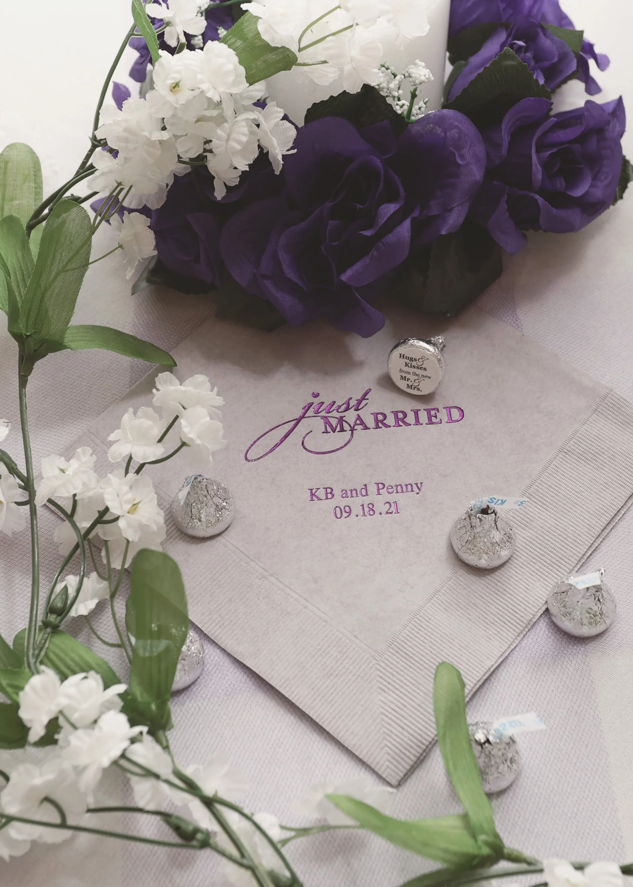 A wedding keepsake with white and purple flowers, a beige cloth with a 'Just Married' inscription, and chocolates wrapped in silver foil and labeled with 'Hugs & Kisses' and 'from the newly Mr. & Mrs.' for KB and Penny's wedding on 09.18.21.
