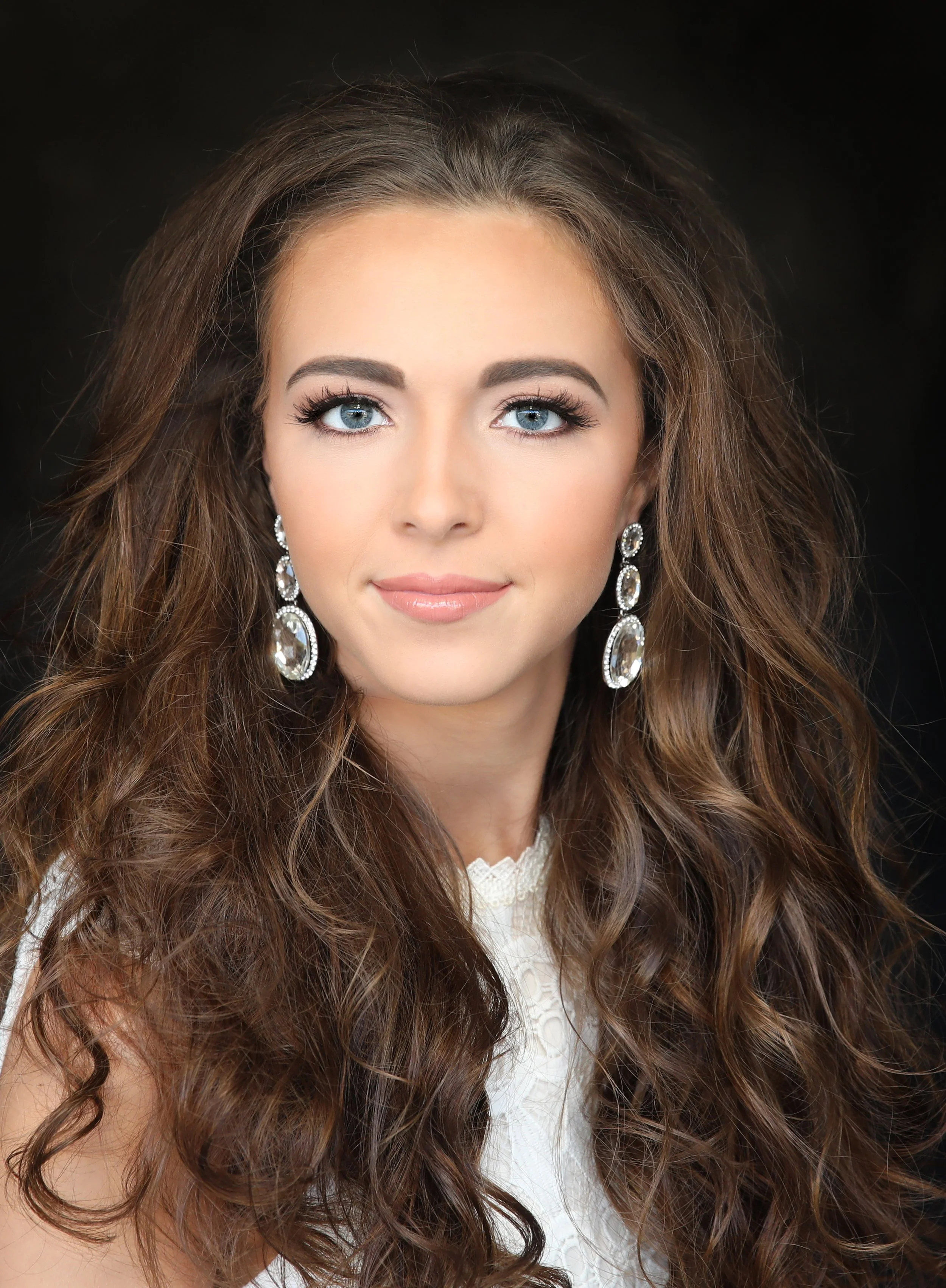 A close-up portrait of a young woman with long, curly brown hair, blue eyes, wearing large sparkling earrings and a white lace top, against a black background.