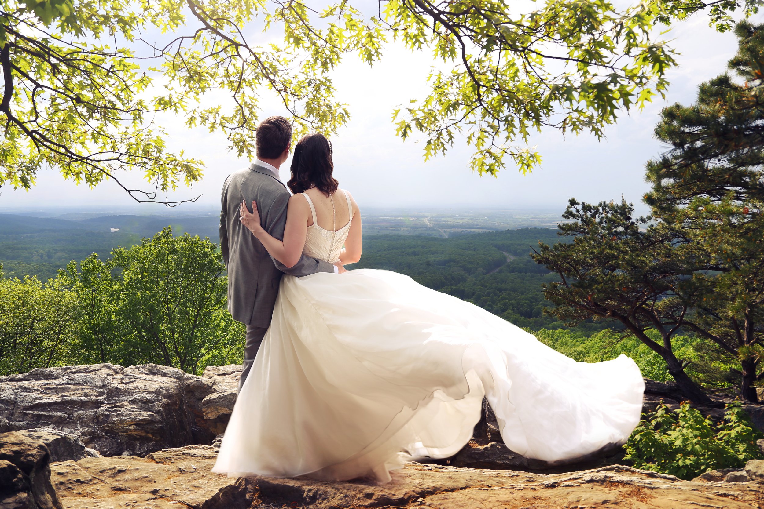 A newlywed couple standing on a rocky ledge, overlooking a forest and distant horizon, with the bride in a white gown and the groom in a gray suit.