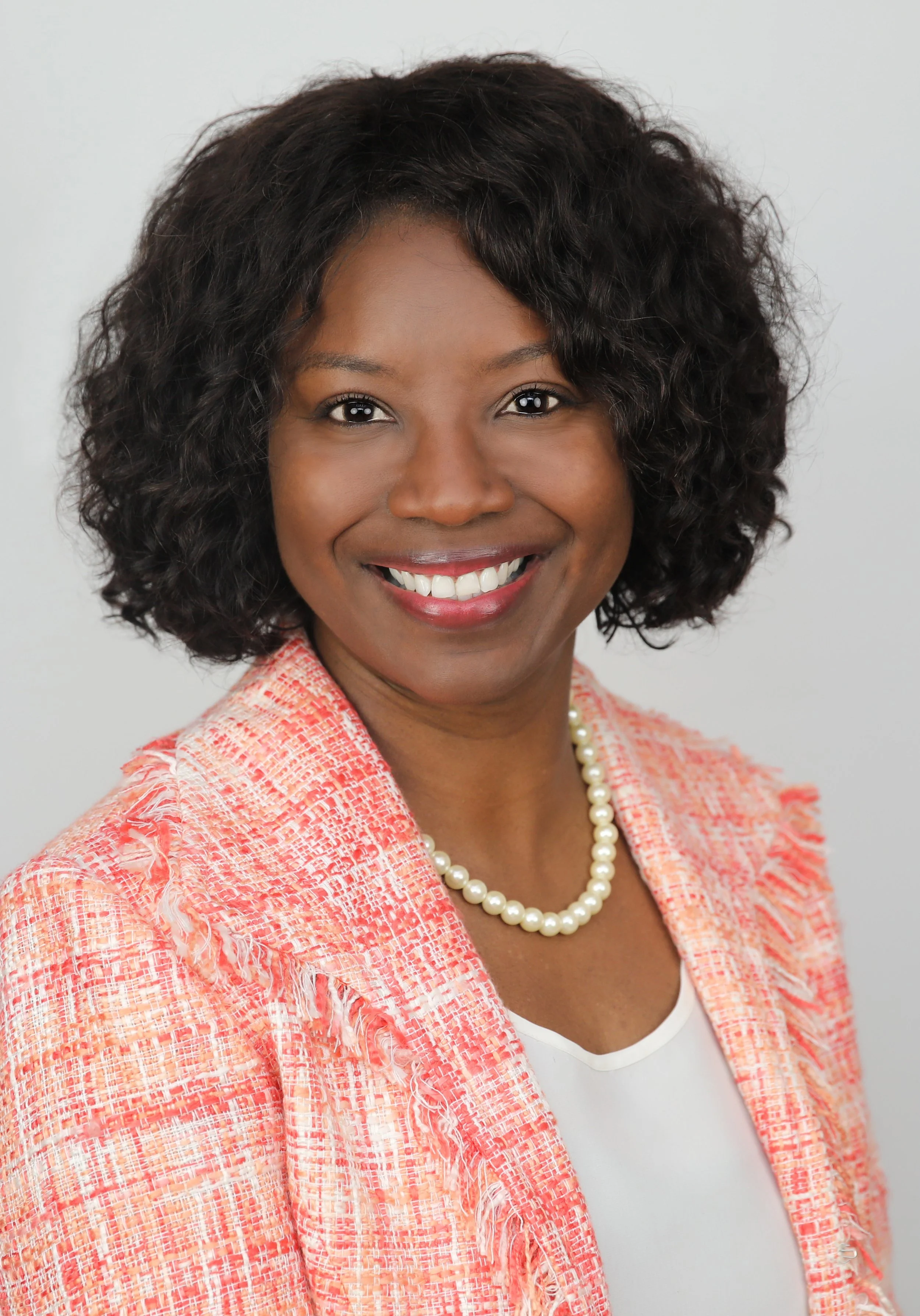 Portrait of a smiling African American woman with curly black hair, wearing a coral textured blazer, a white top, and a pearl necklace, against a plain light gray background.