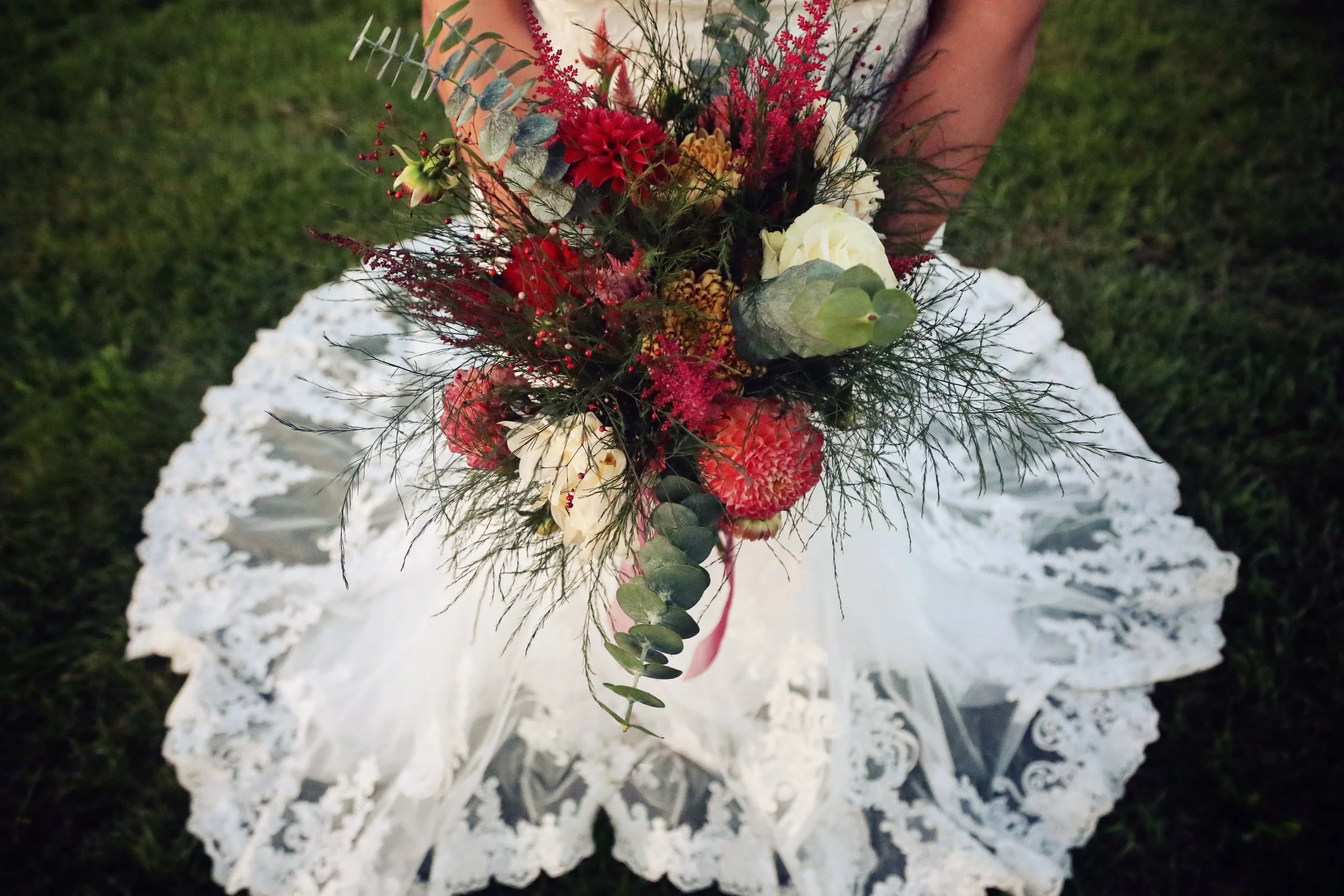 A person in a white lace dress holding a bouquet of red, white, and green flowers and foliage, with grass in the background.