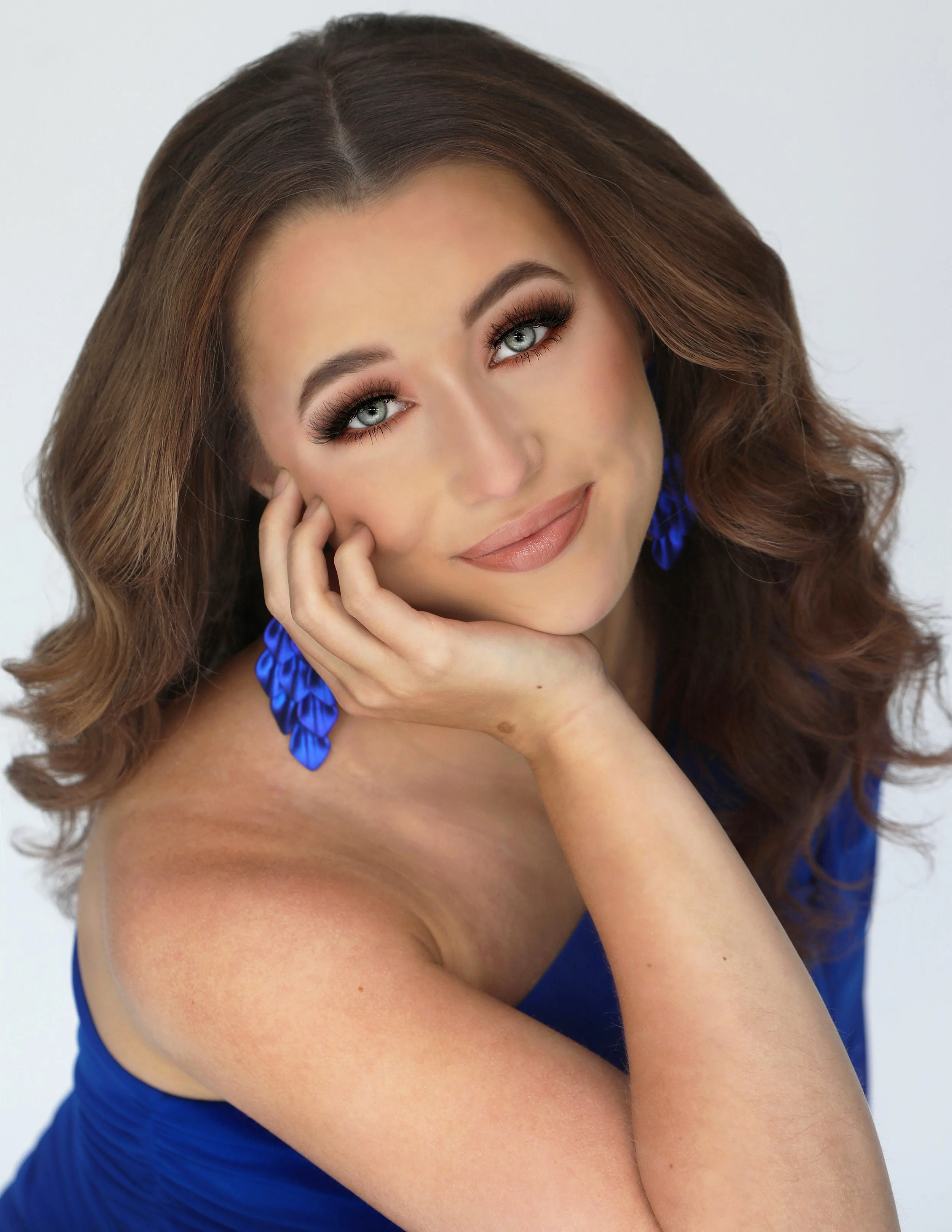 A woman with long brown wavy hair and blue eyes, wearing blue earrings and a matching blue dress, resting her head on her hand and smiling at the camera.