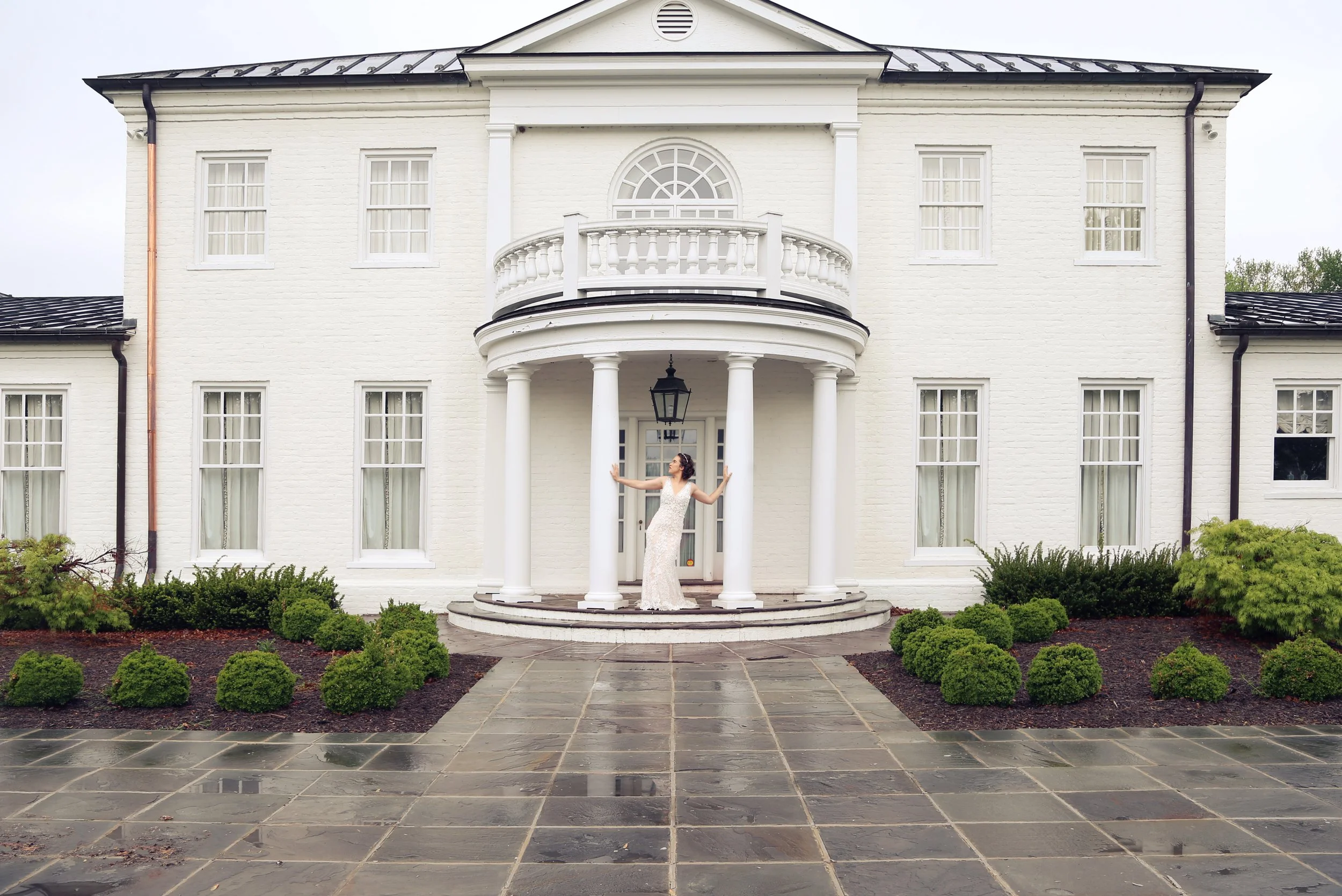 A woman in a wedding dress standing on the steps of a white mansion's porch, surrounded by landscaping and wet stone pavement.
