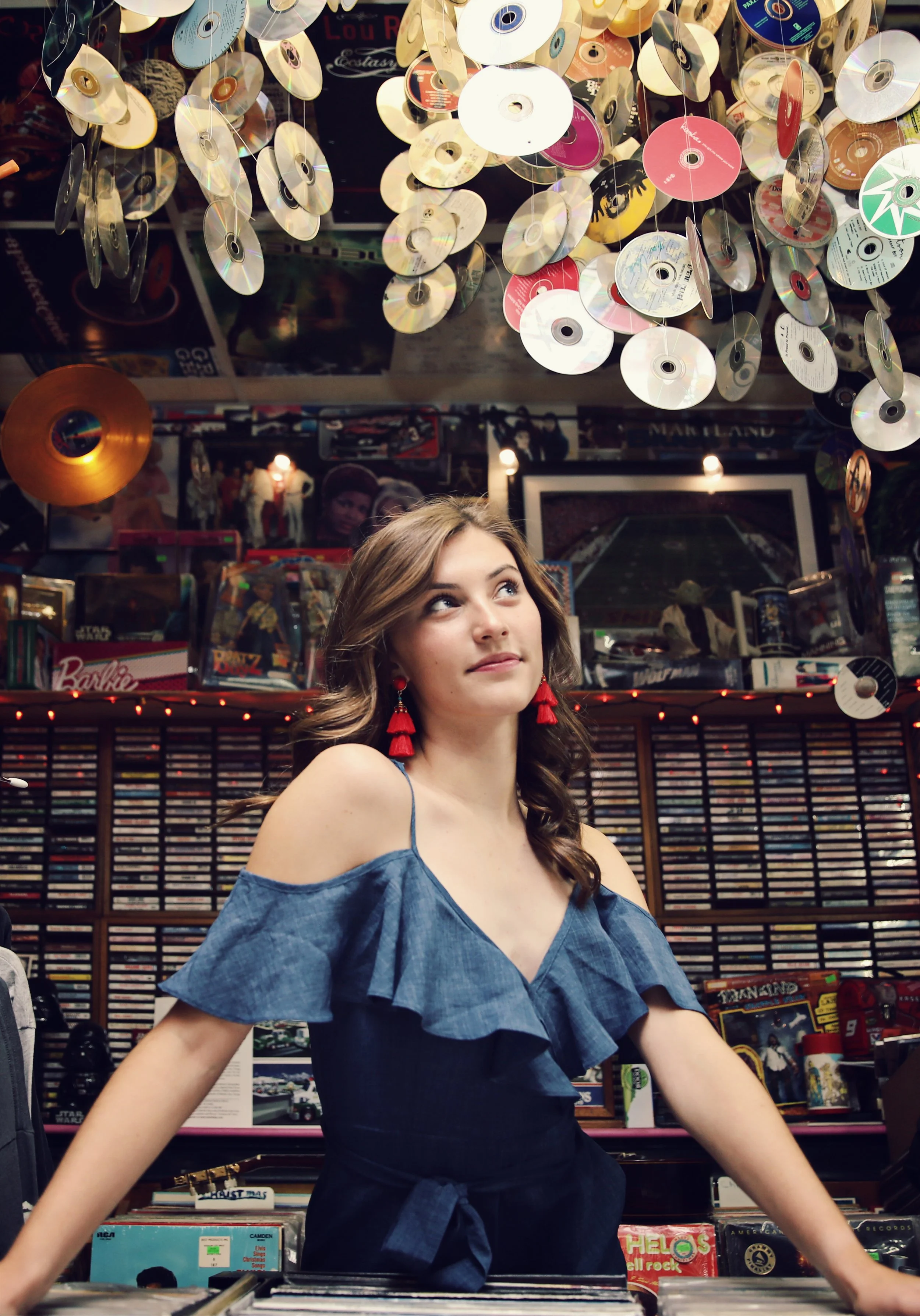 Young woman with brown hair wearing red tassel earrings and a denim off-shoulder ruffled top in a music store surrounded by CDs and vinyl records, with hanging CDs from the ceiling.