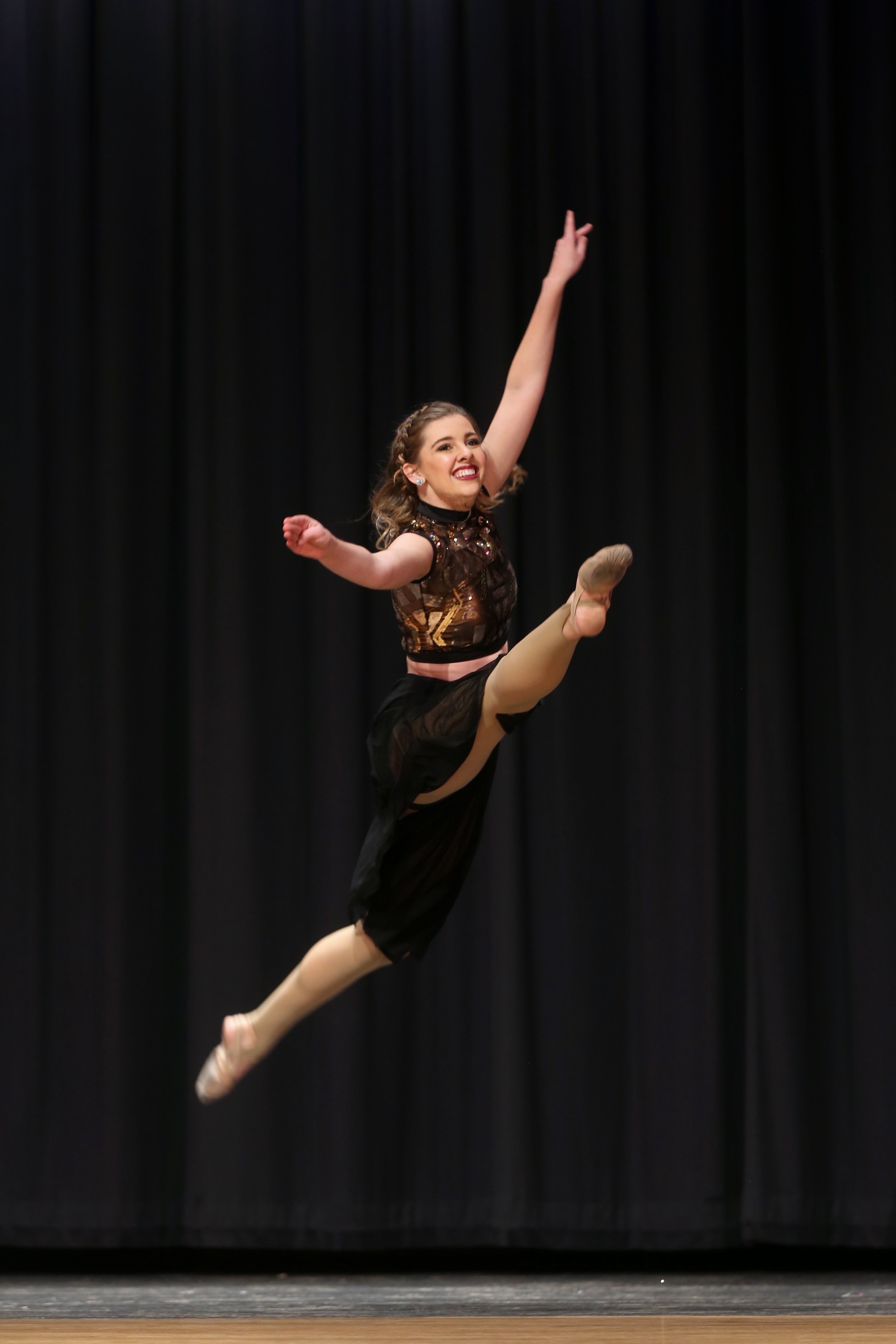 A young woman performing an energetic dance leap on stage, wearing a black and gold costume, with a black curtain background.