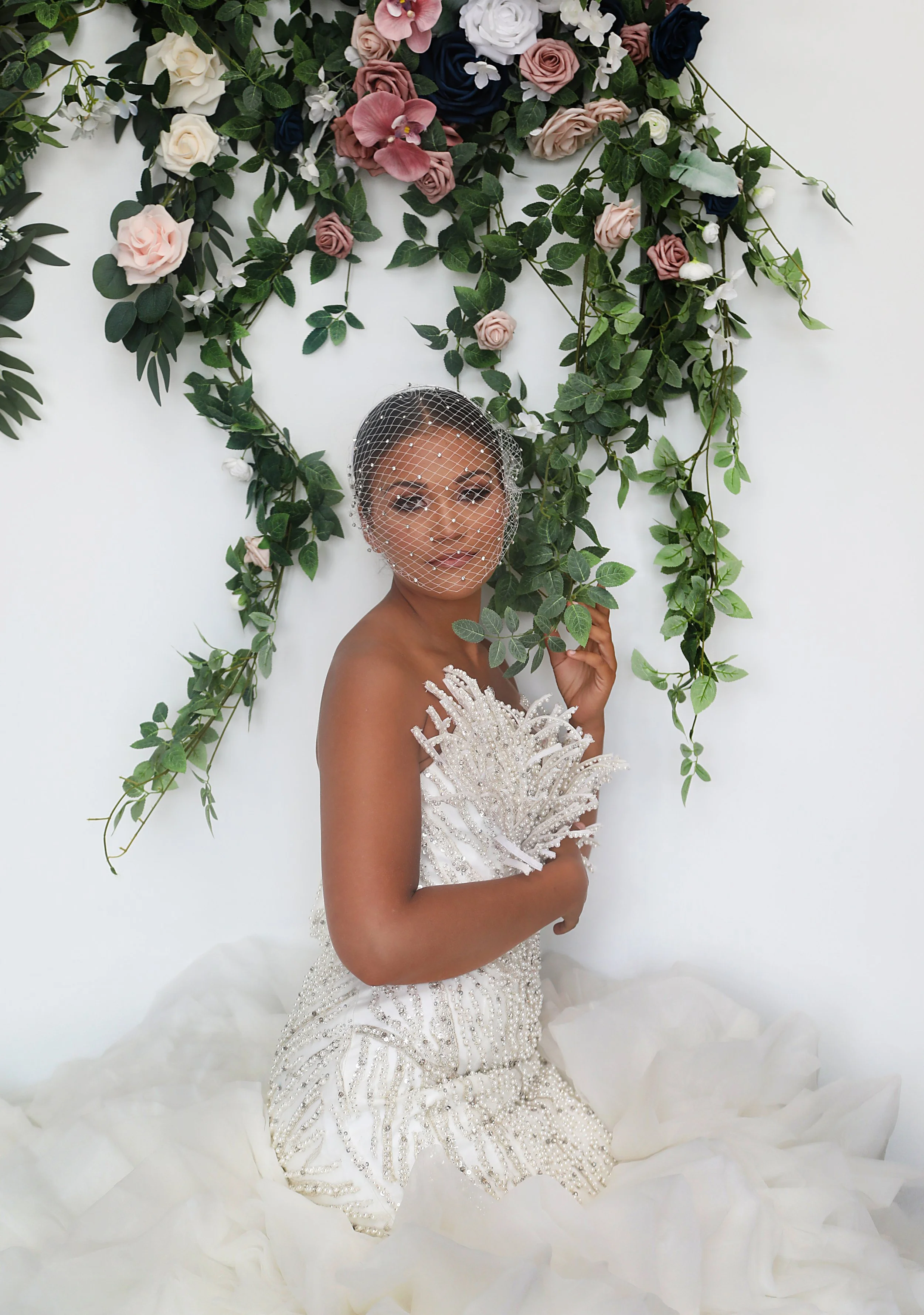 Bridal portrait of a woman in an ornate white gown with beads, wearing a birdcage veil, standing in front of a floral backdrop with pink, white, and dark blue roses and green leaves.