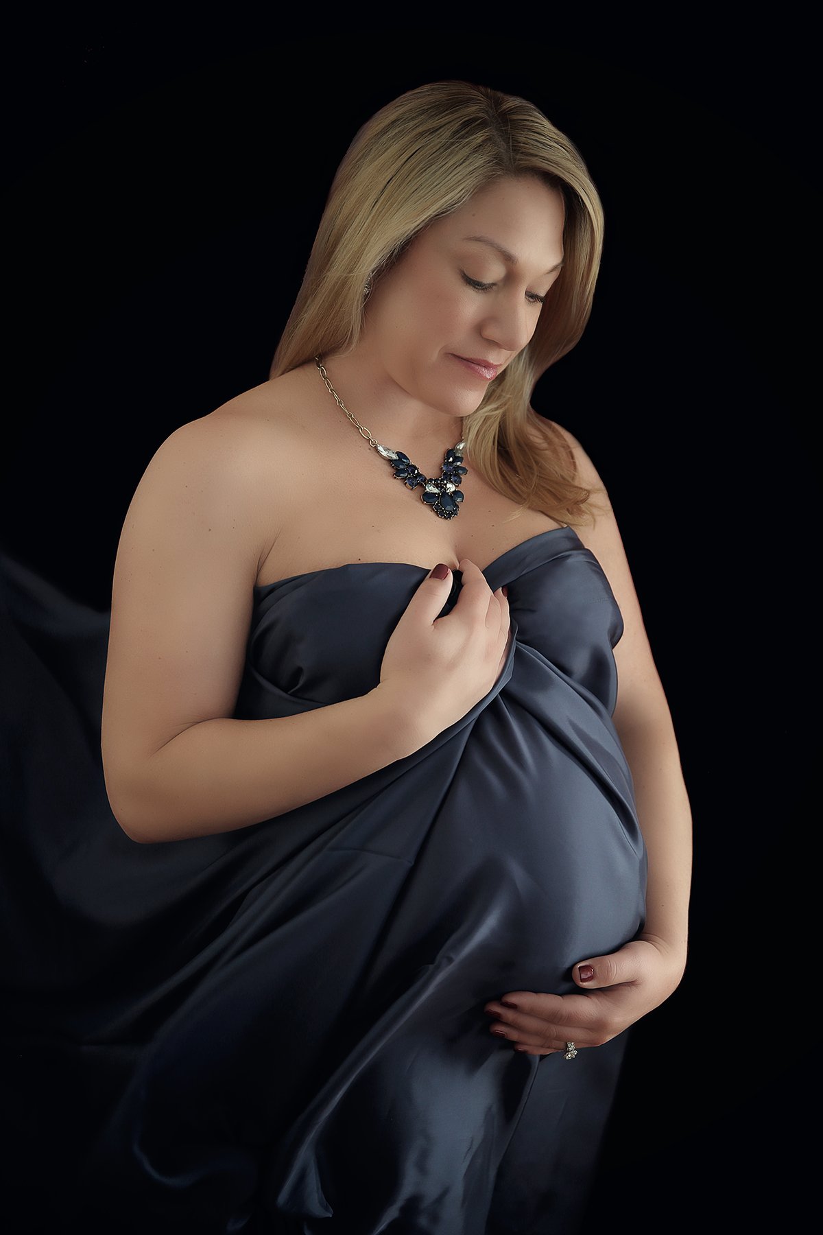 A pregnant woman in a strapless dark satin dress, wearing a navy necklace, gently holding her belly and her chest, looking downward against a black background.