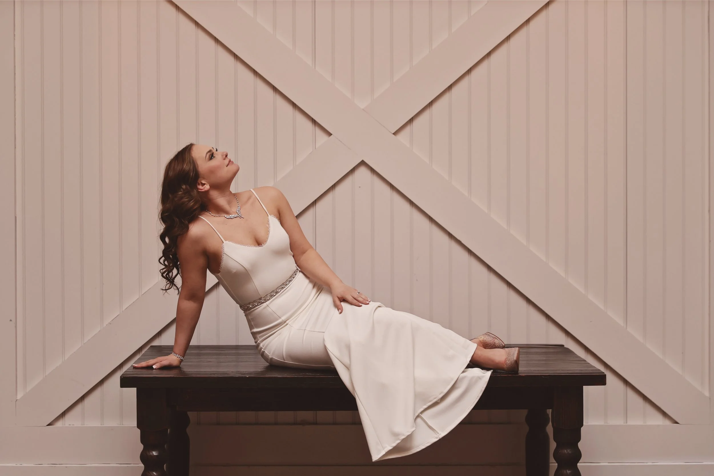 A woman in a white dress sitting on a dark wooden bench against a beige paneled wall with white cross beams.