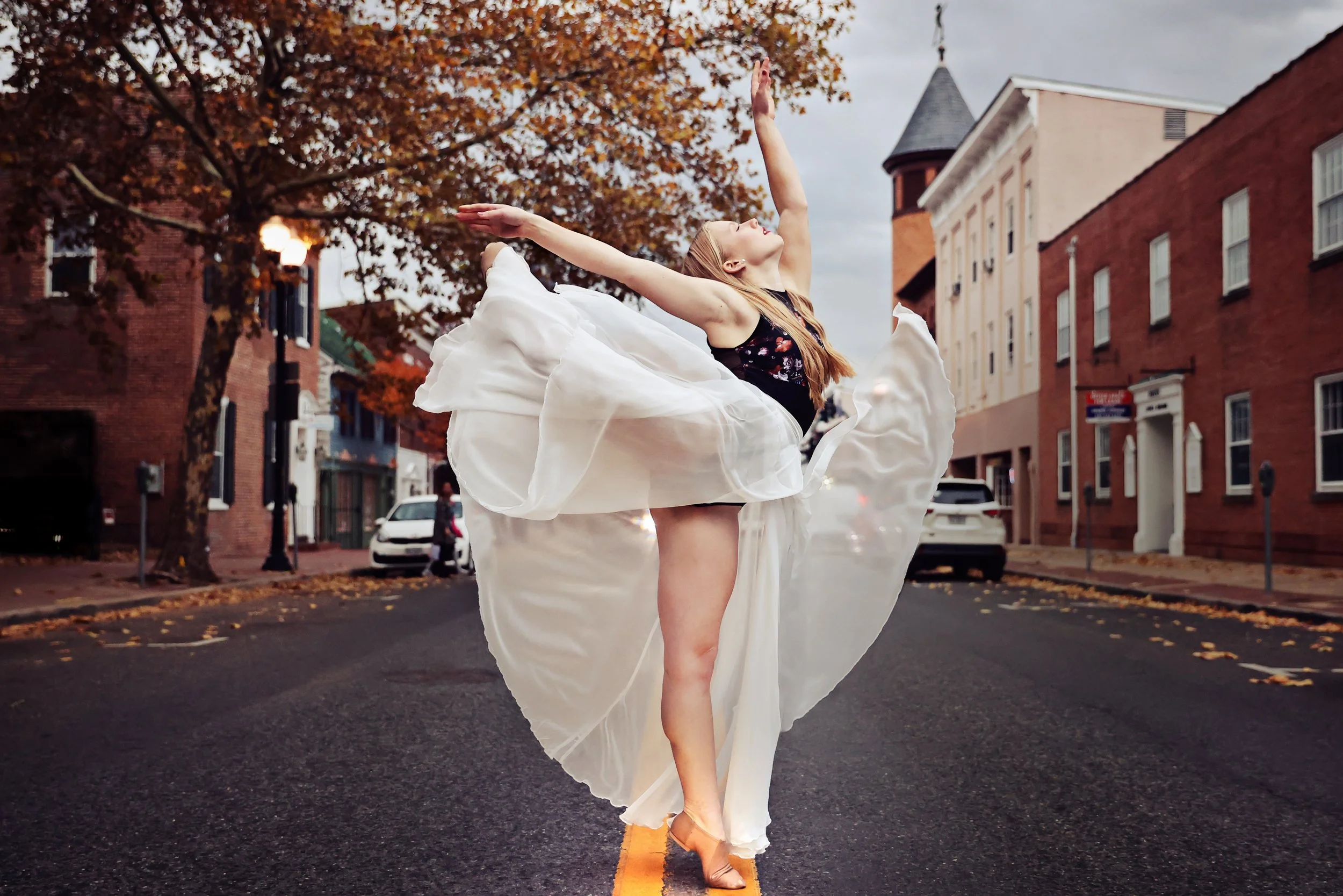 A ballet dancer performing on an empty city street with brick buildings and trees with fall foliage in the background.