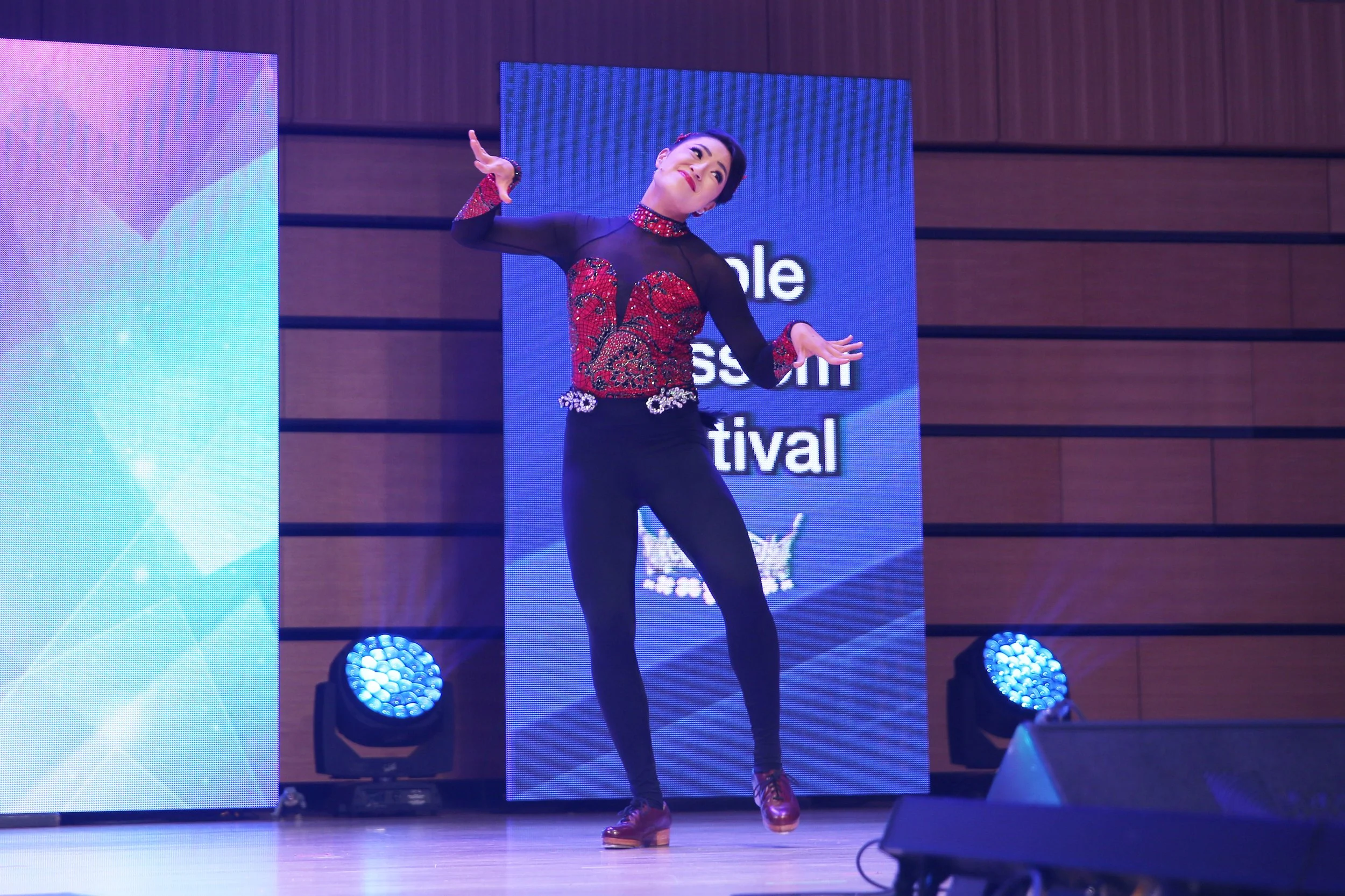 A woman performing a dance on stage at the Singapore Asian Festival, wearing a black and red embellished dance costume and brown shoes.