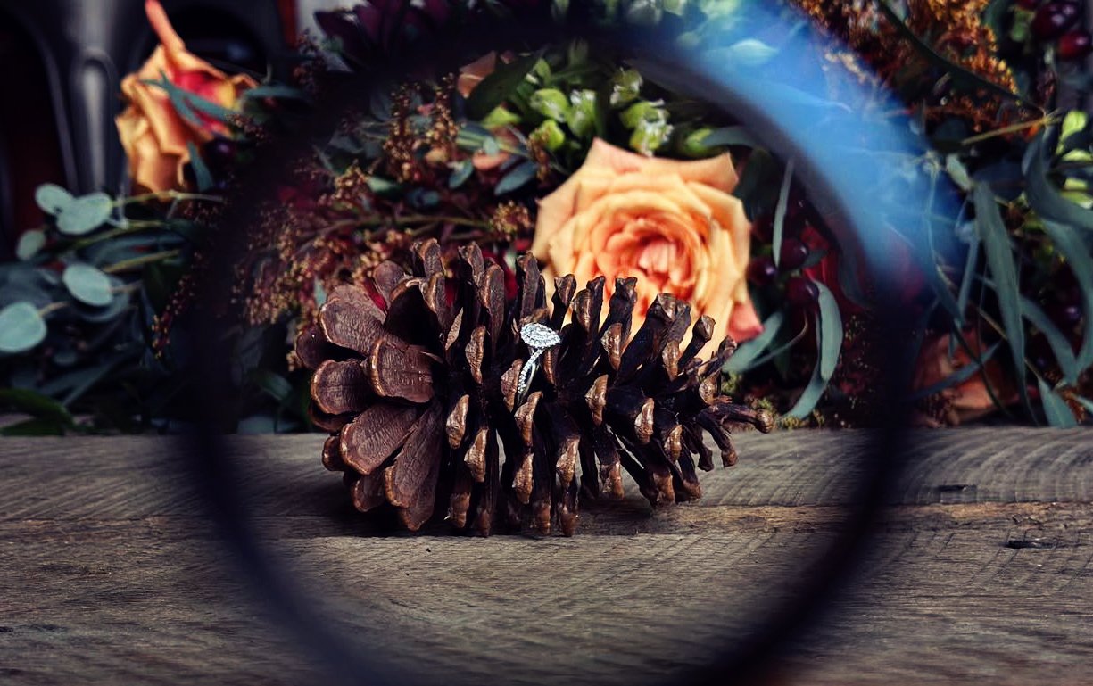 Pinecone on a wooden surface with a blurred bouquet of flowers and greenery in the background.